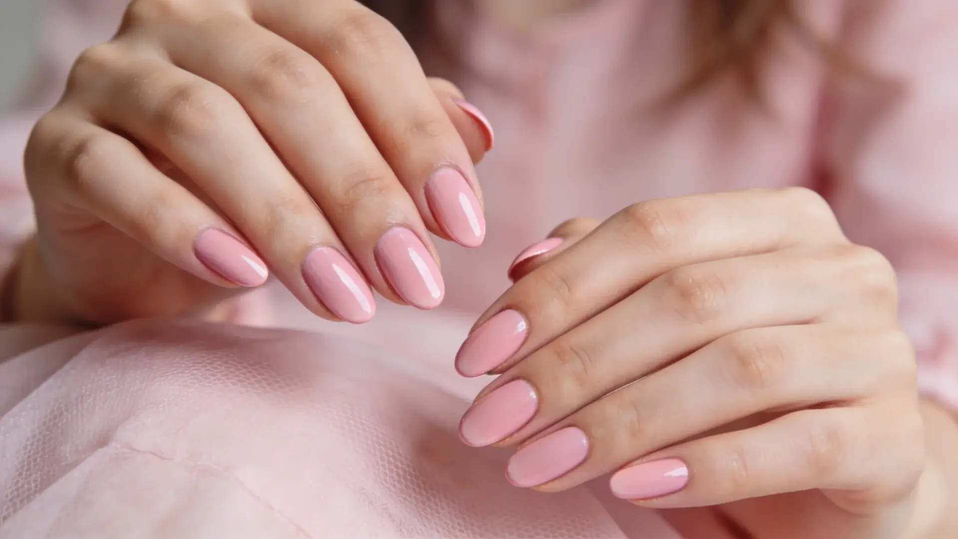 Glossy baby pink manicure with short oval nails on hands placed on soft pink fabric
