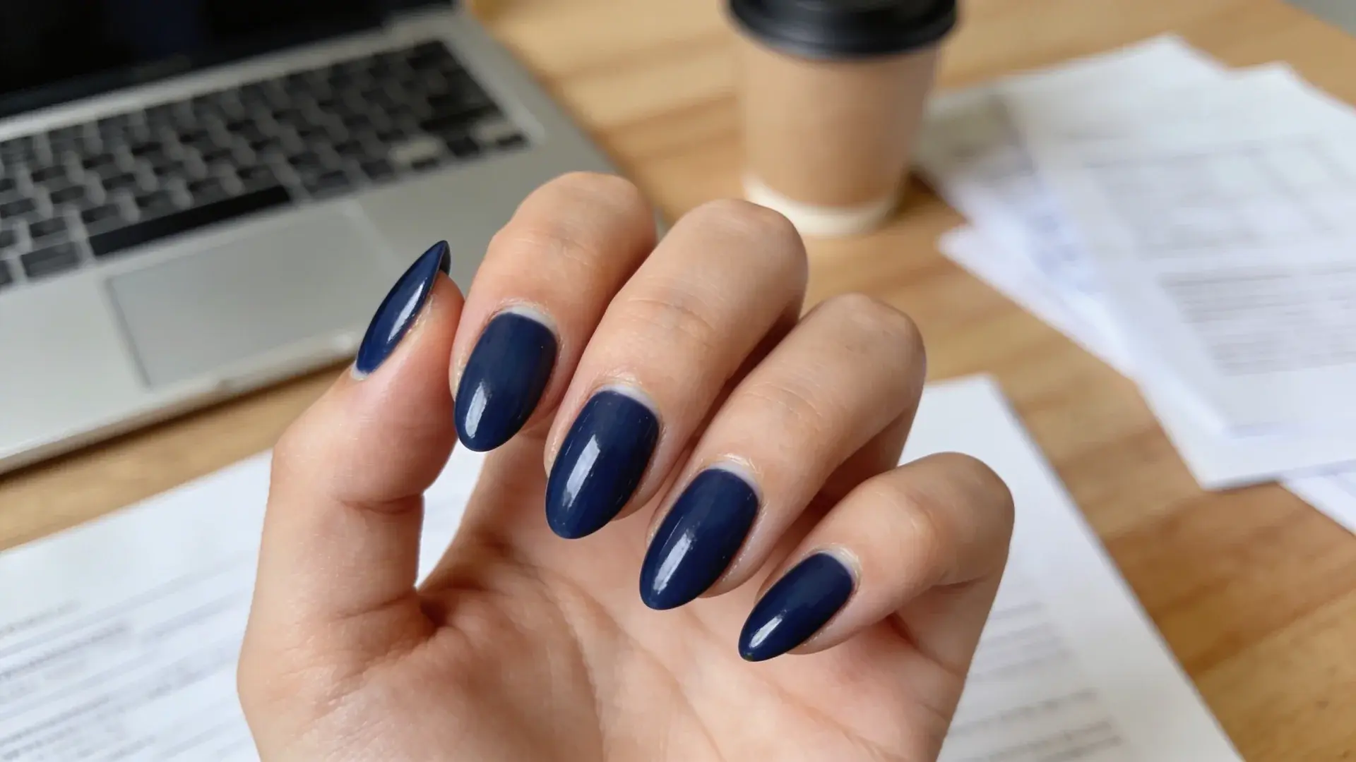Glossy navy blue almond manicure shown near laptop and coffee cup on office desk