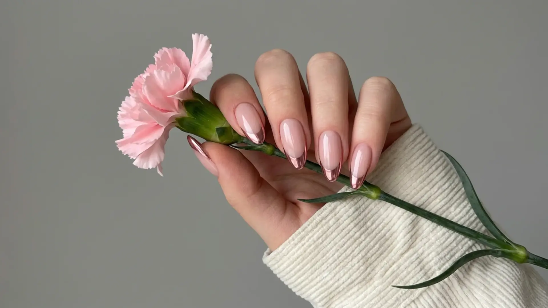 Hand holding pink flower with glossy nude manicure and metallic rose gold chrome French tips on almond nails
