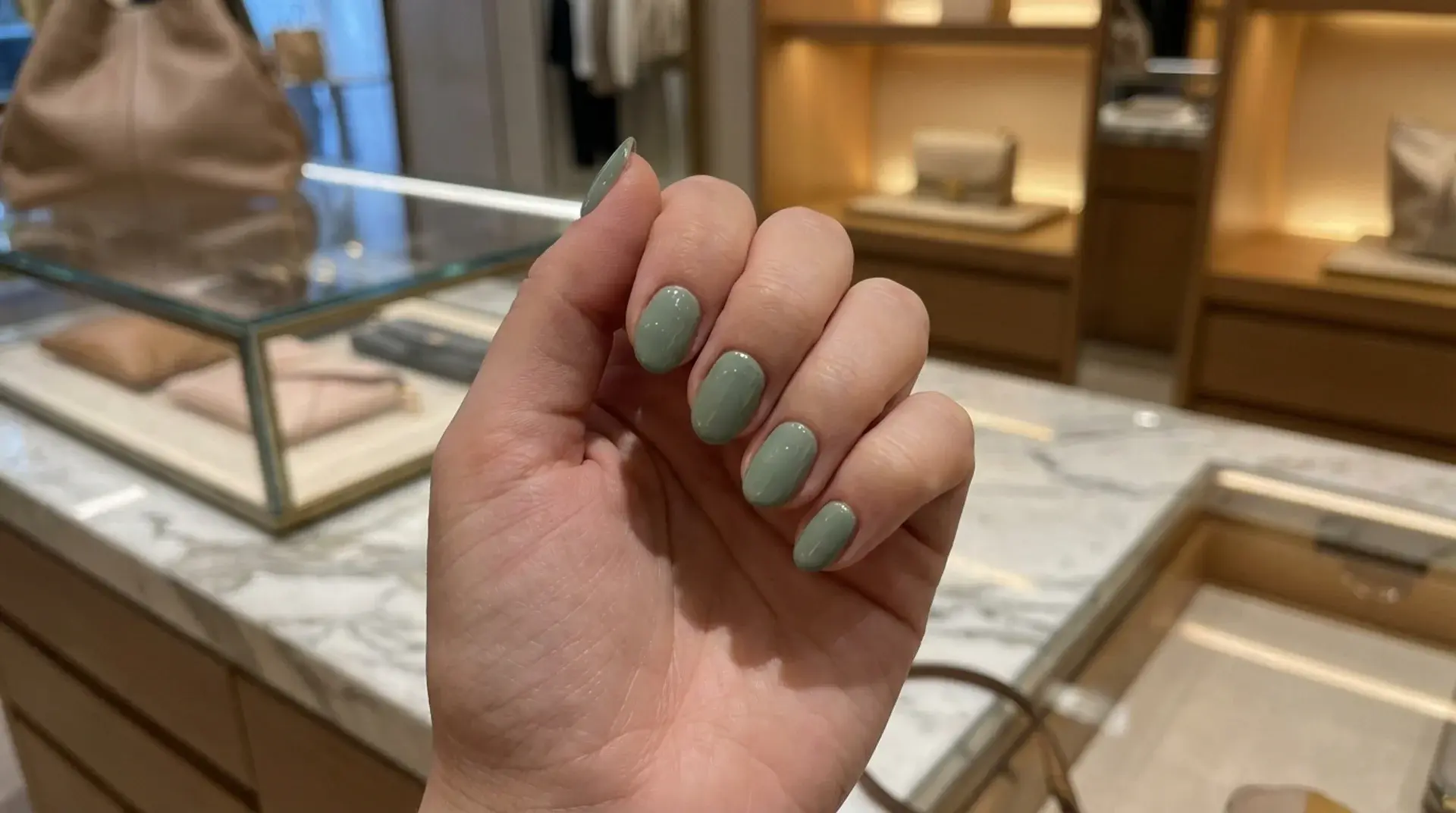 Hand showing glossy sage green manicure with almond shaped nails inside luxury boutique with marble counter display