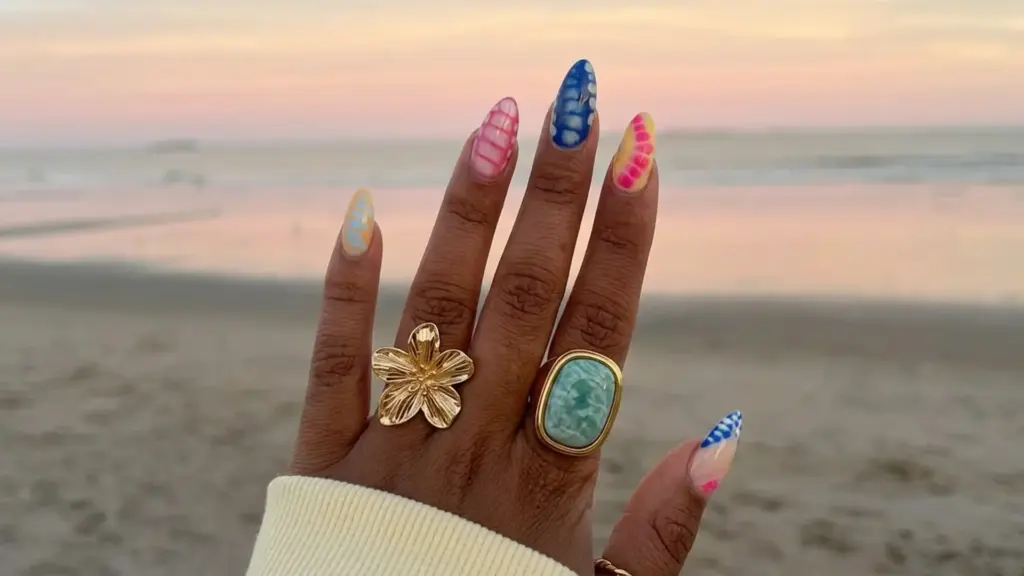 Hand with colorful patterned manicure and gold rings held up at beach sunset showing vibrant spring break nails design