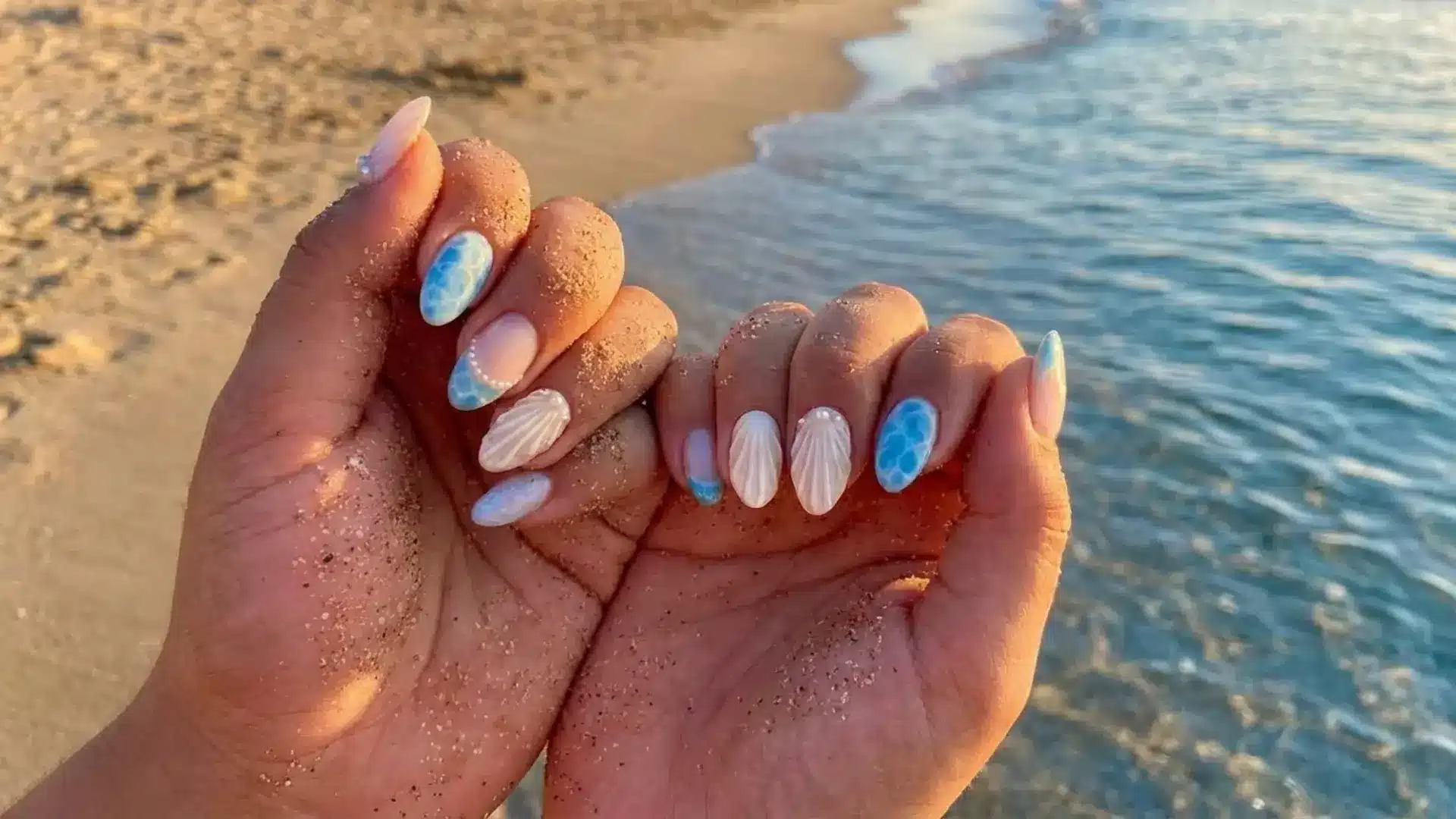 Hands with sandy fingers at beach showing glossy ocean themed manicure with shells and pearls showing 3D accent beach nails design