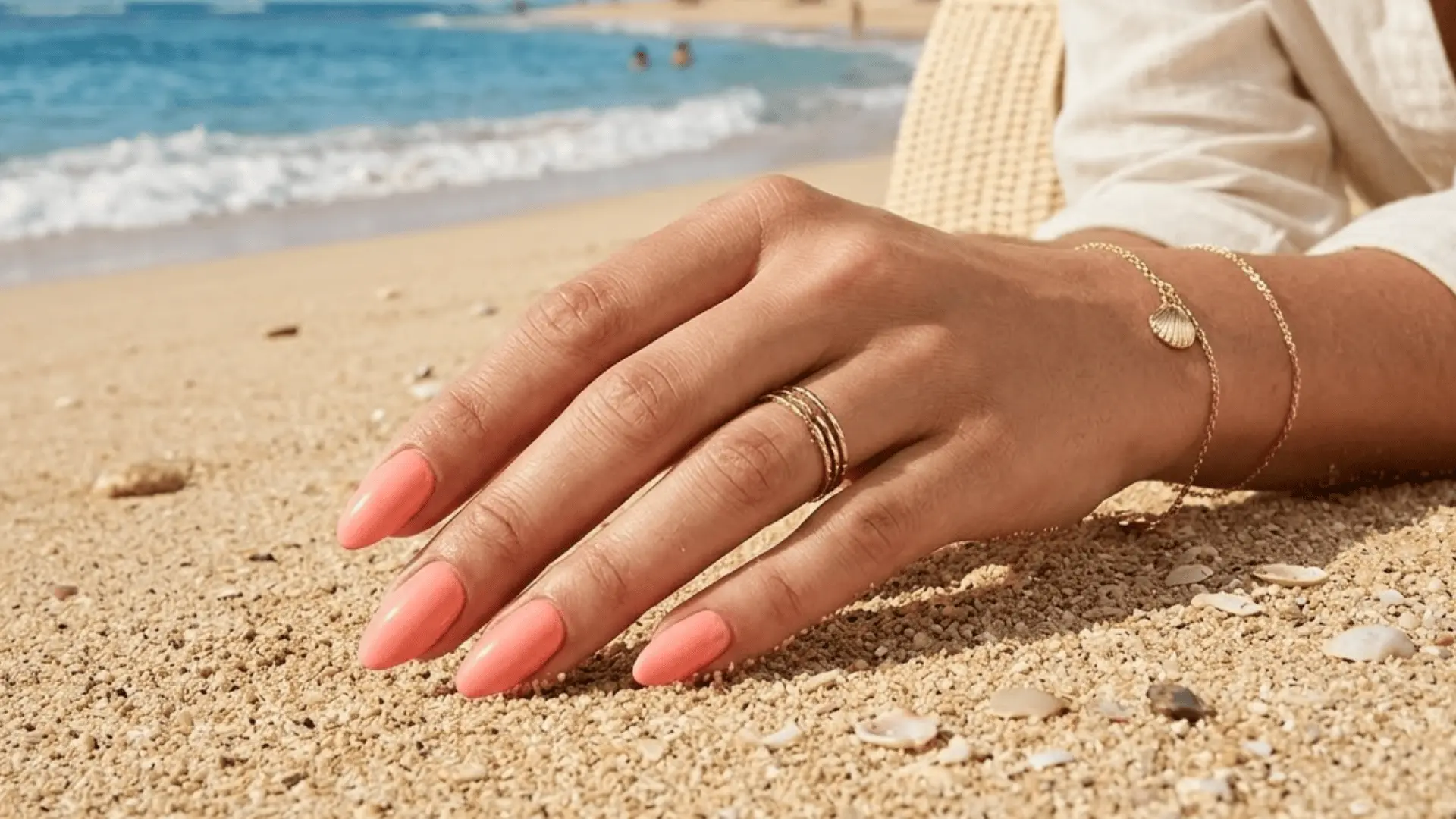 Peachy coral manicure resting on sandy beach with ocean waves in the background