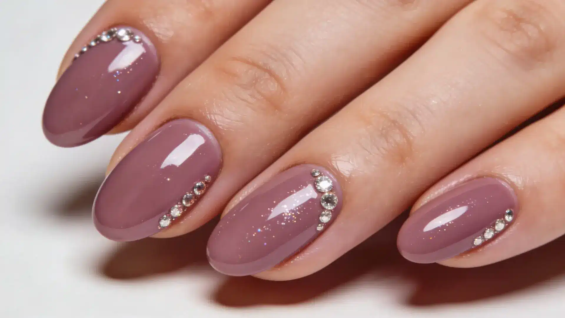 Small rhinestones placed near the cuticle or along one side add sparkle to mauve color nails. on a small oval shape nails