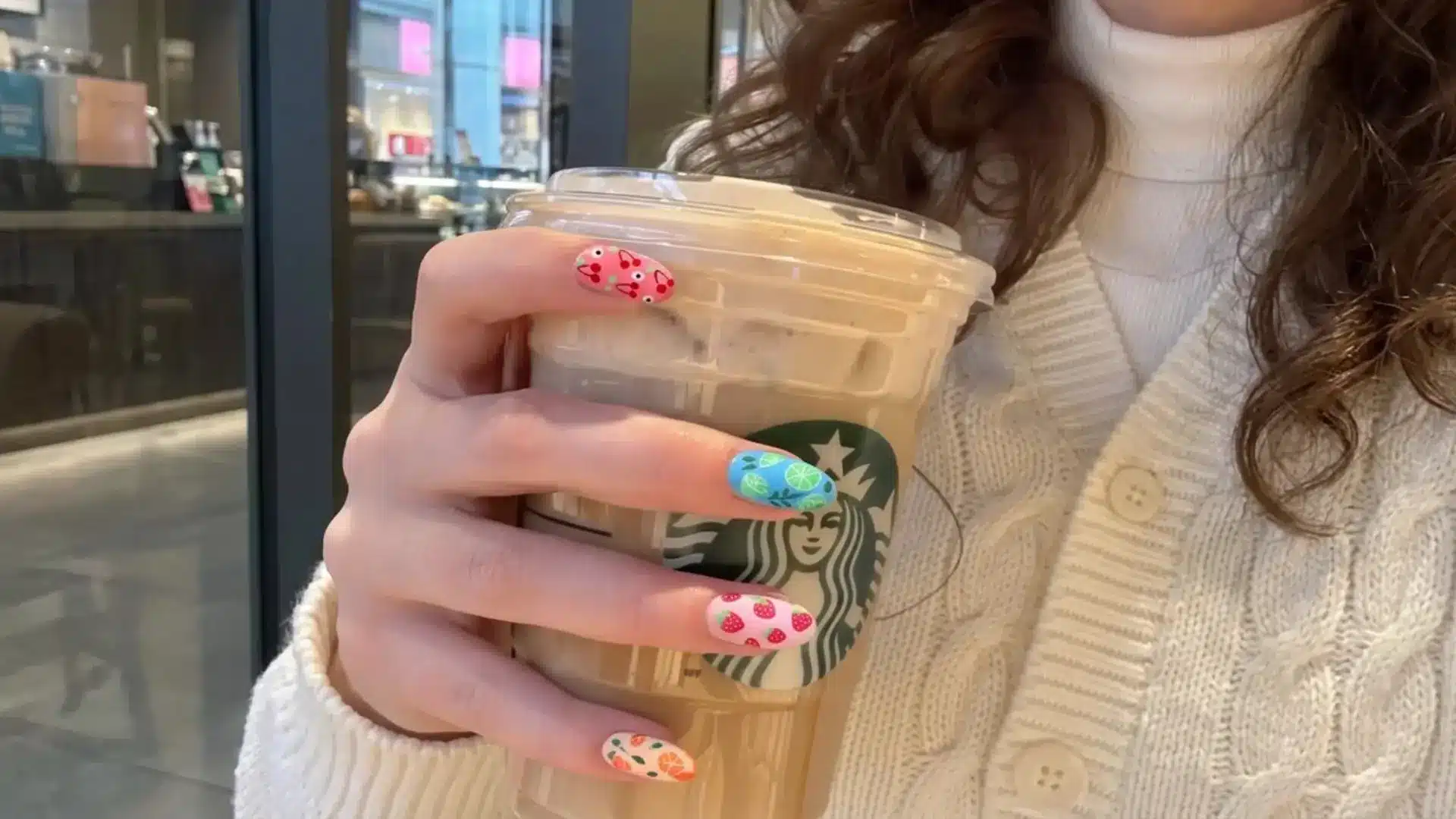 Spring break nails idea showing colorful fruit nail art manicure holding Starbucks iced coffee cup wearing a white knit sweater