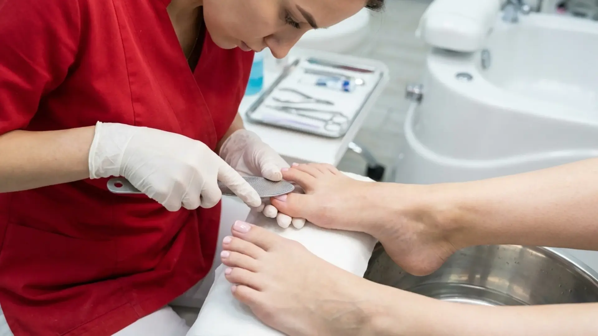 Technician in red uniform filing toenails during pedicure treatment with tools and foot basin in a salon