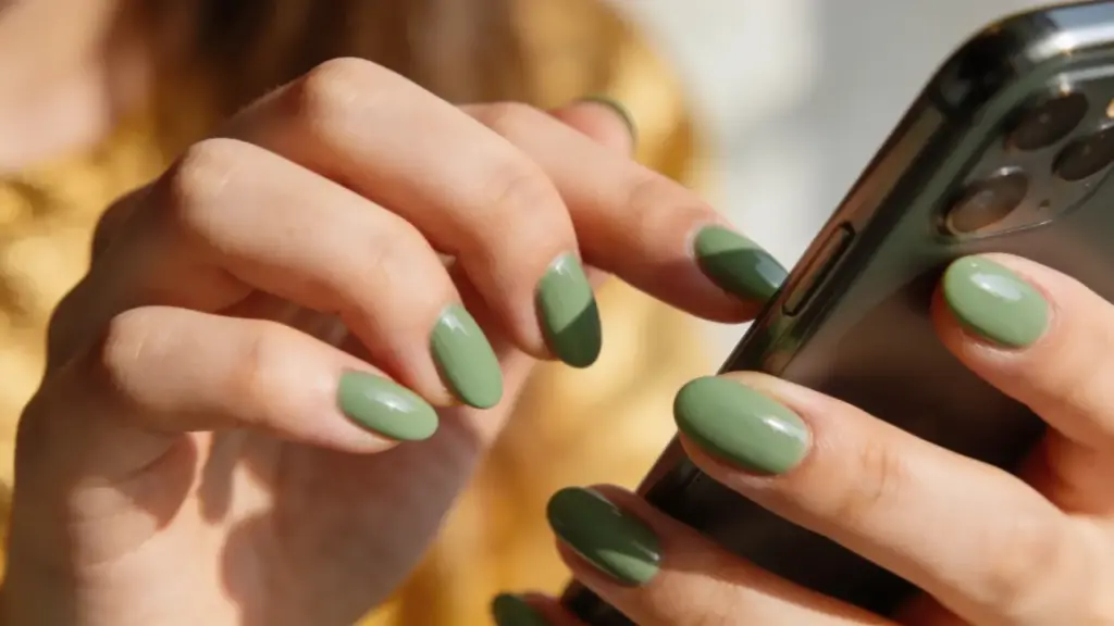 Woman holding smartphone with glossy sage green manicure and almond shaped nails in warm natural sunlight