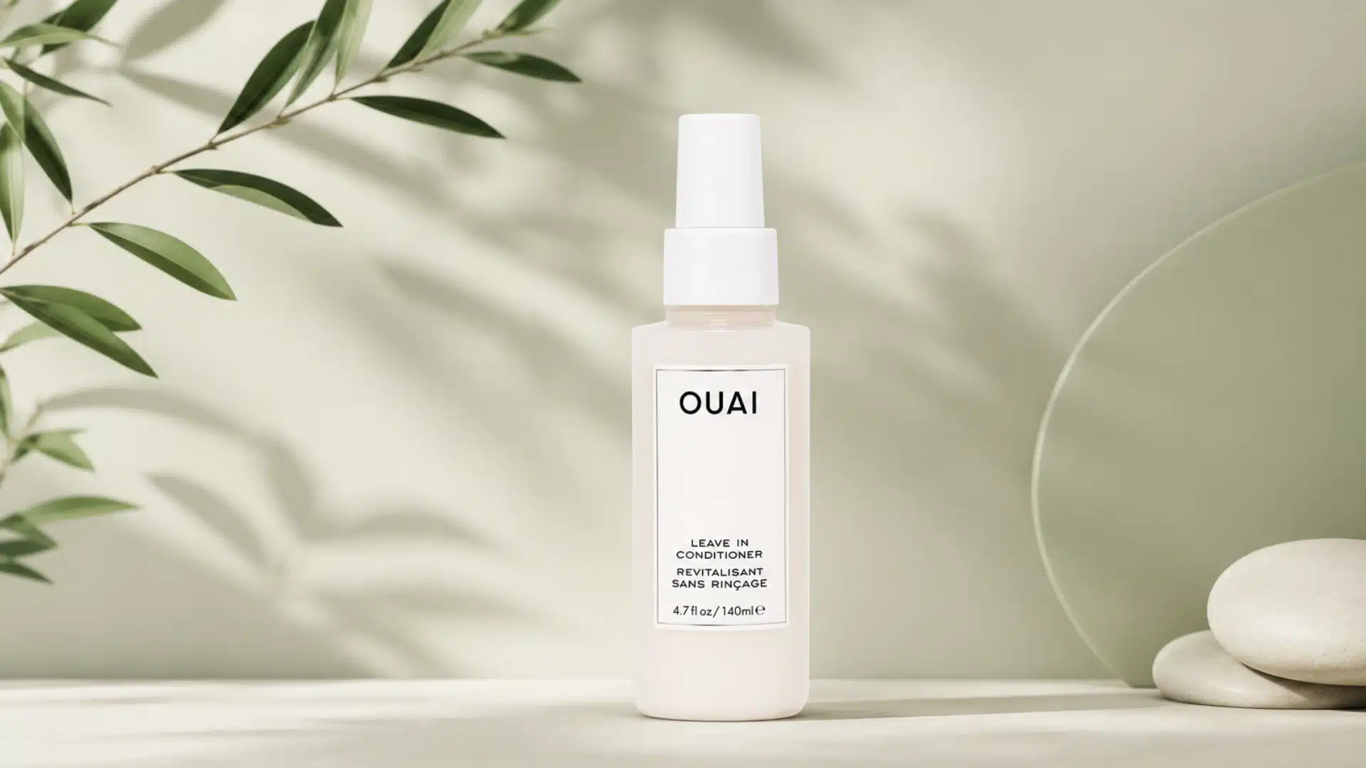 A bottle of OUAI Leave In Conditioner centered against a soft green background with leafy shadows and smooth stones