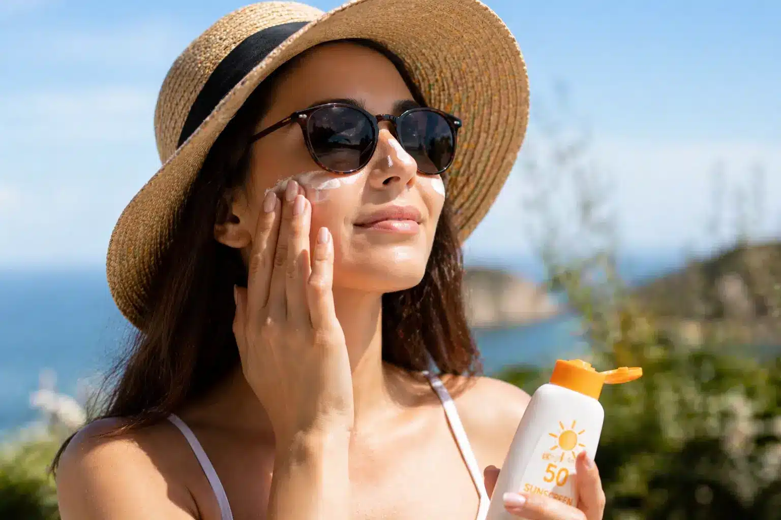 A smiling woman in a sun hat and sunglasses applies SPF 50 sunscreen to her face at a sunny coastal location.