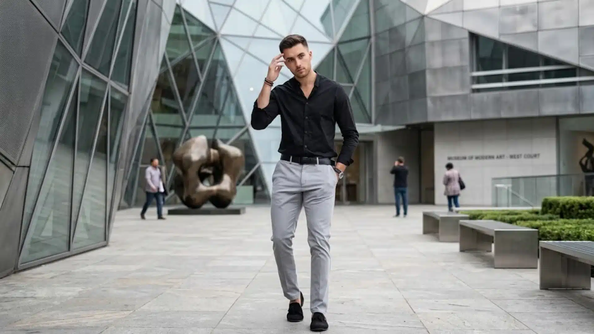 A stylish man in a black button-down and grey trousers poses in front of a modern glass art museum