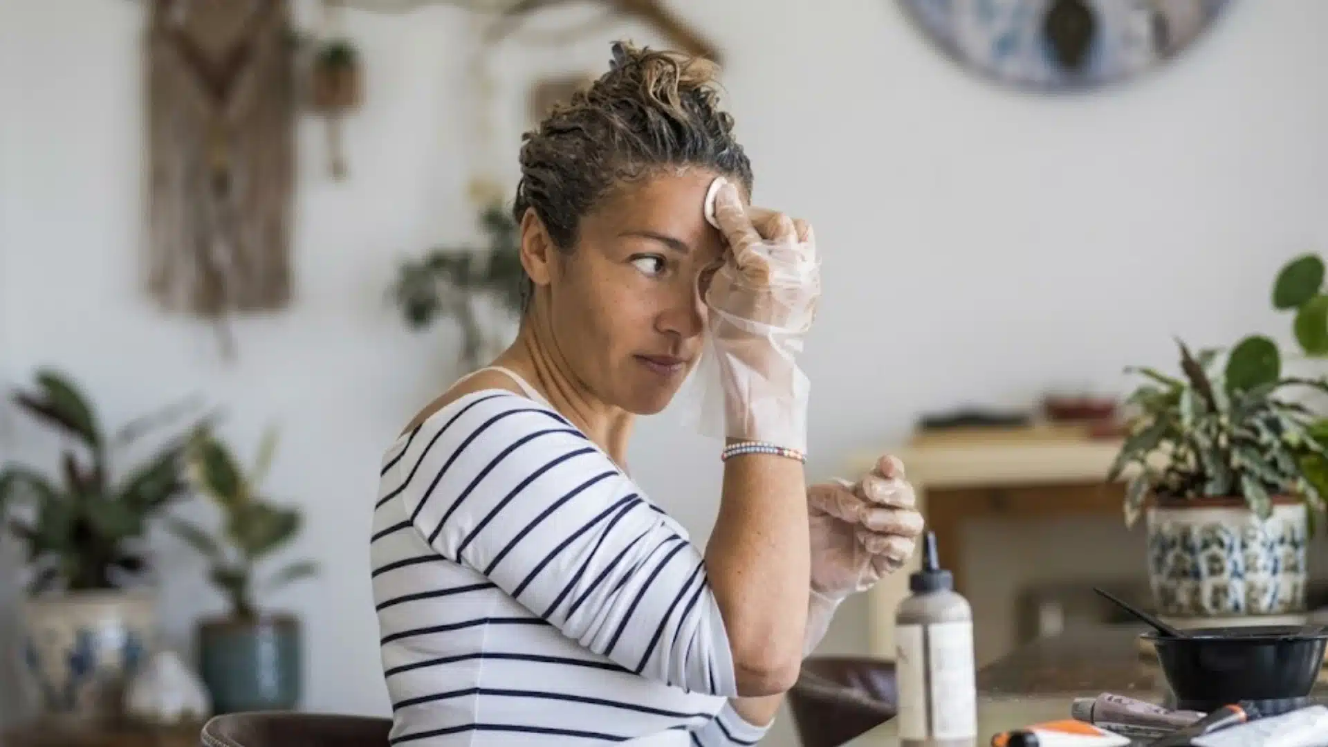 A woman in a striped shirt, wearing gloves, wipes hair dye from her forehead while seated at a home table