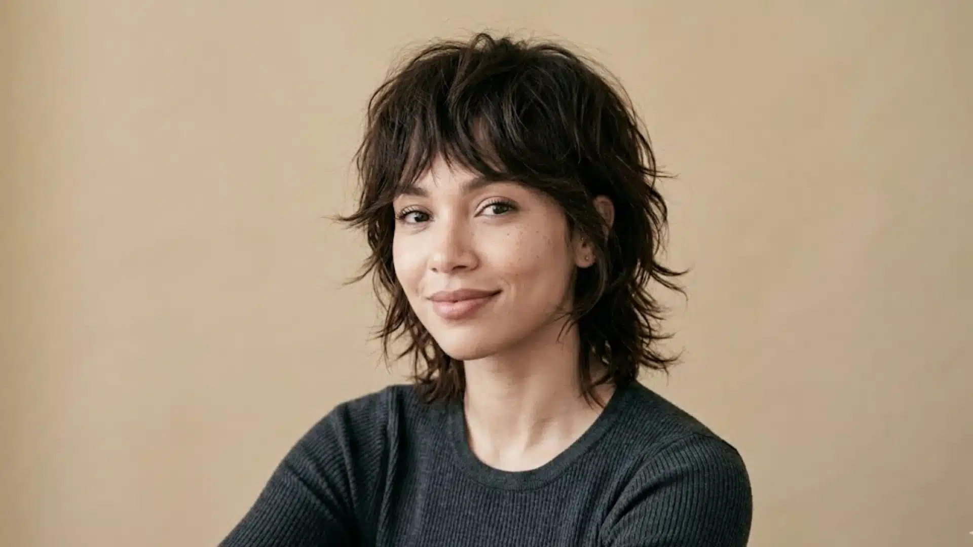 A woman with a textured, brunette shag haircut smiling against a plain beige background