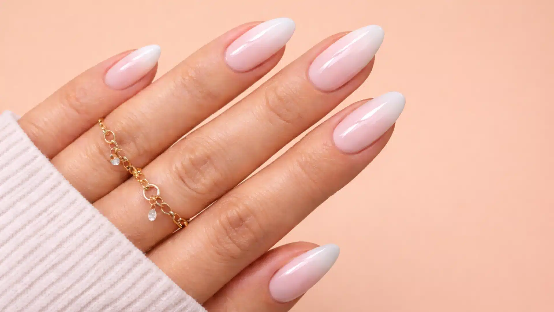 Almond-shaped soft pink ombre nails with glossy finish and gold rings, set against a warm peach solid background
