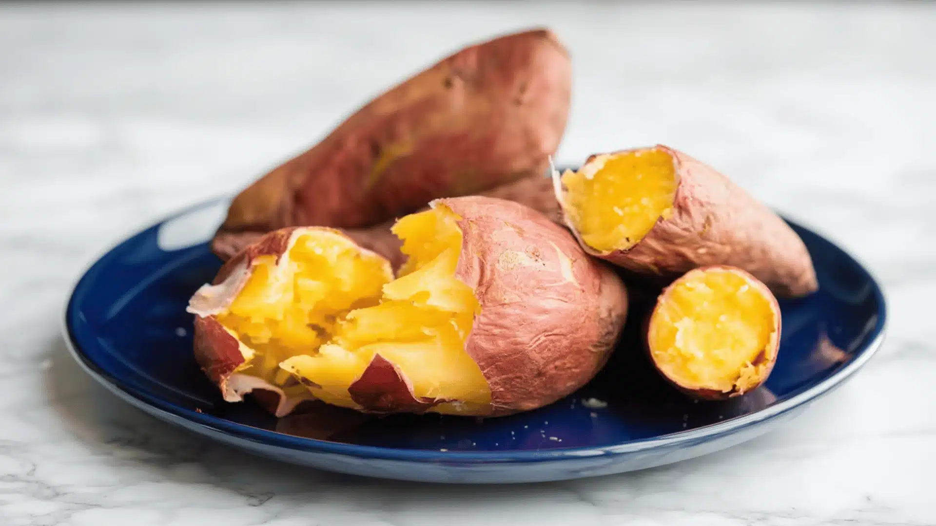 Baked sweet potatoes on a blue plate with soft yellow flesh and wrinkled skin on a marble surface
