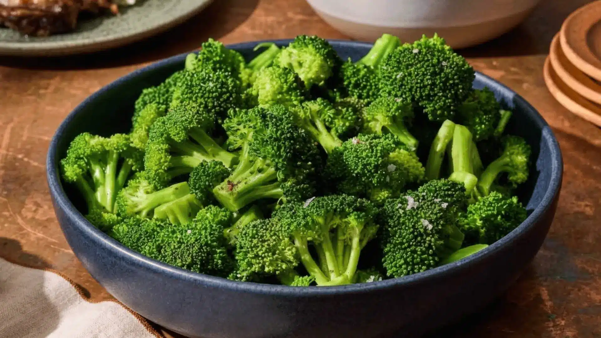 Bowl of fresh steamed broccoli florets seasoned with coarse salt on a wooden dining table