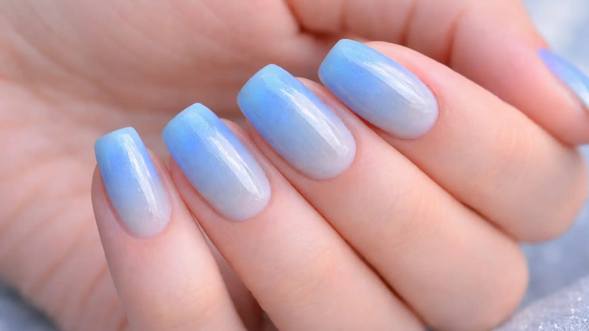 Close-up of a hand with glossy ombre nails in soft white to blue gradient, smooth finish, natural skin tone, blurred background