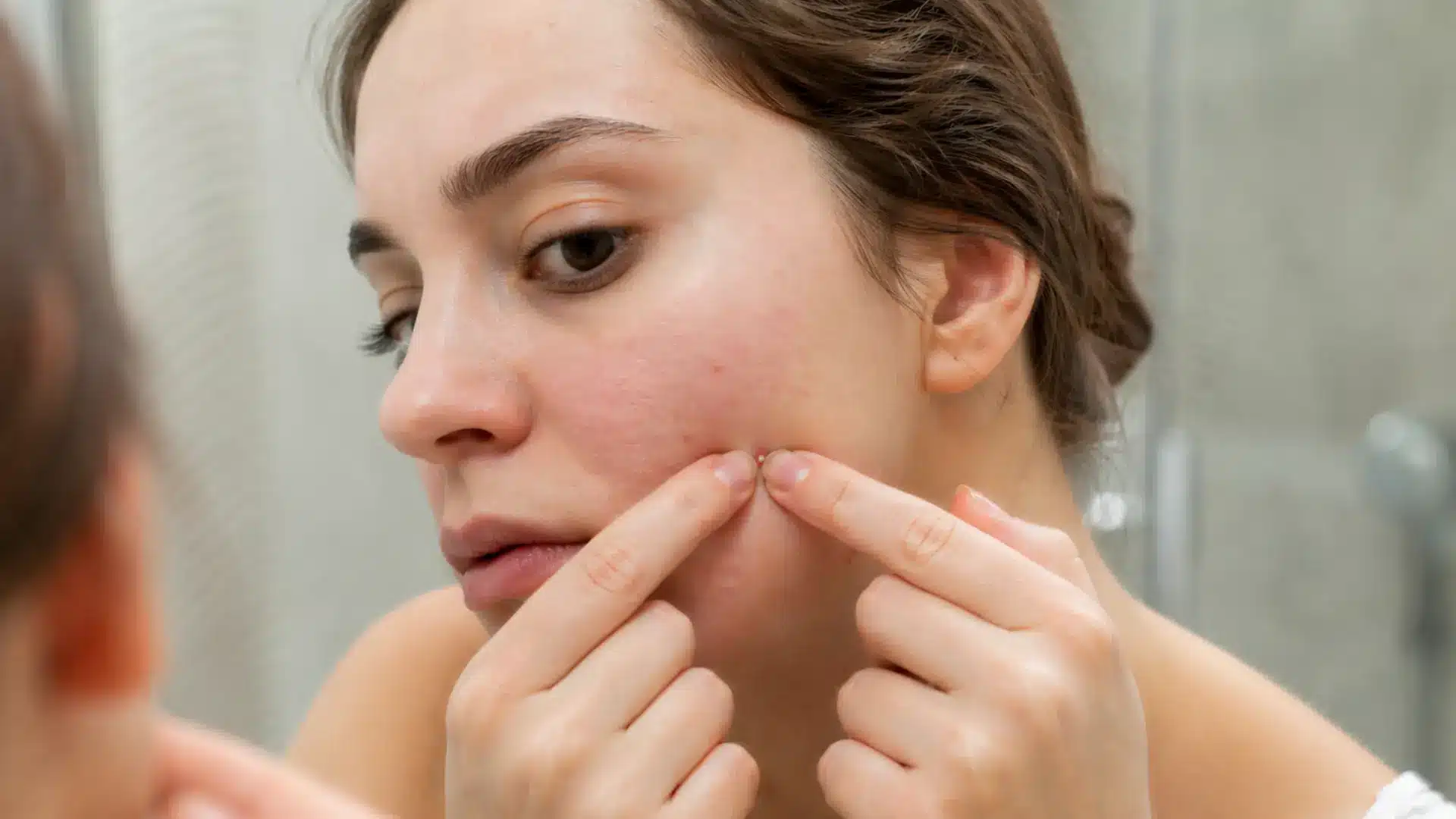 Close-up of a woman squeezing a pimple on her cheek while looking in a mirror