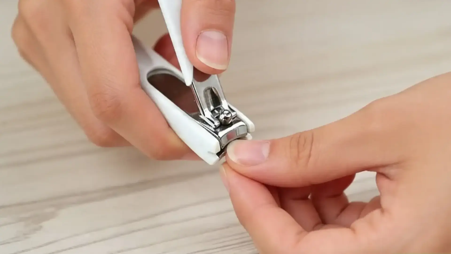 Close-up of hands trimming a fingernail using a white nail clipper on a wooden surface