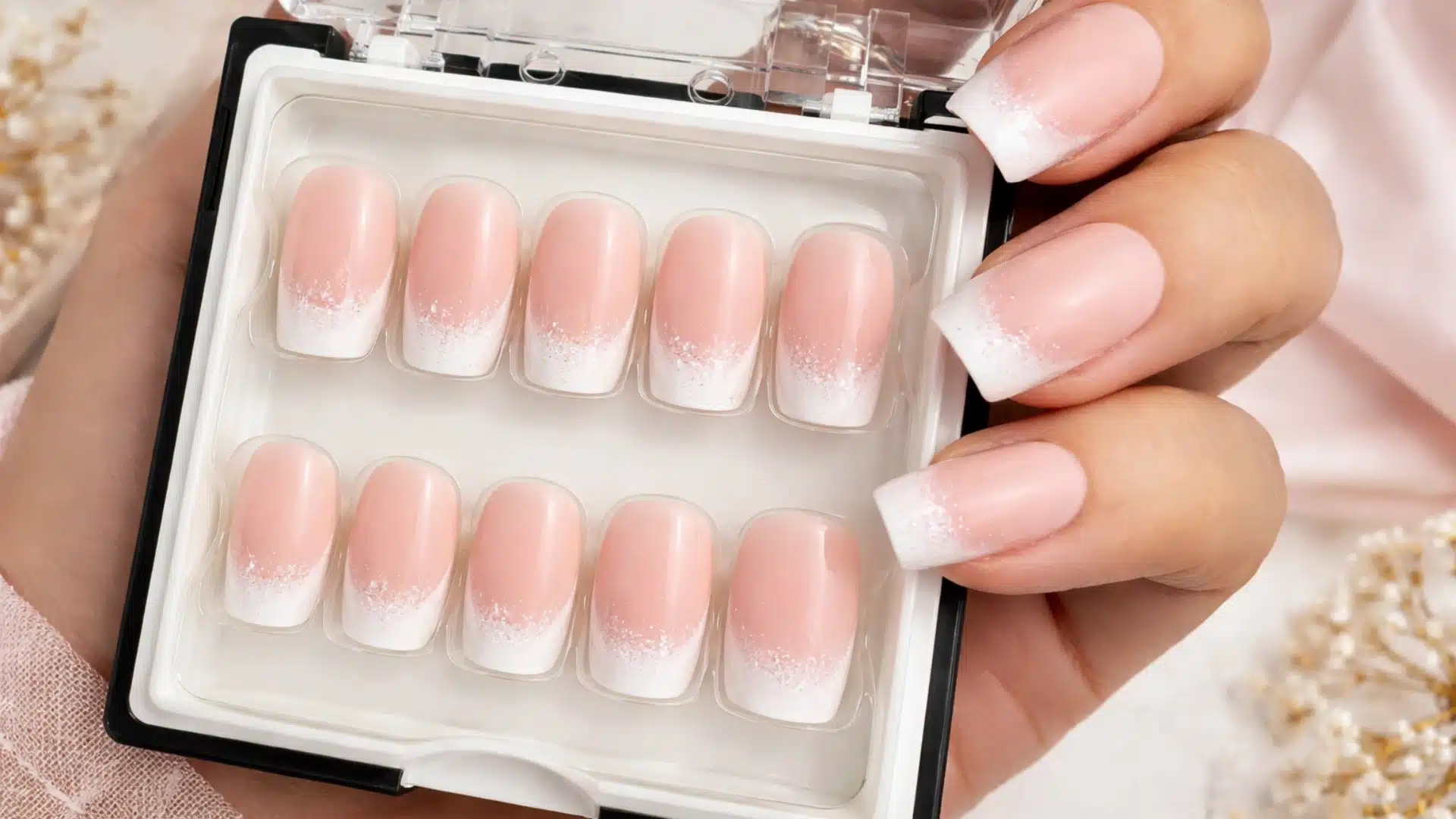 Close-up of press-on nails in a case with soft pink ombre French tips and subtle glitter, held against a warm neutral background