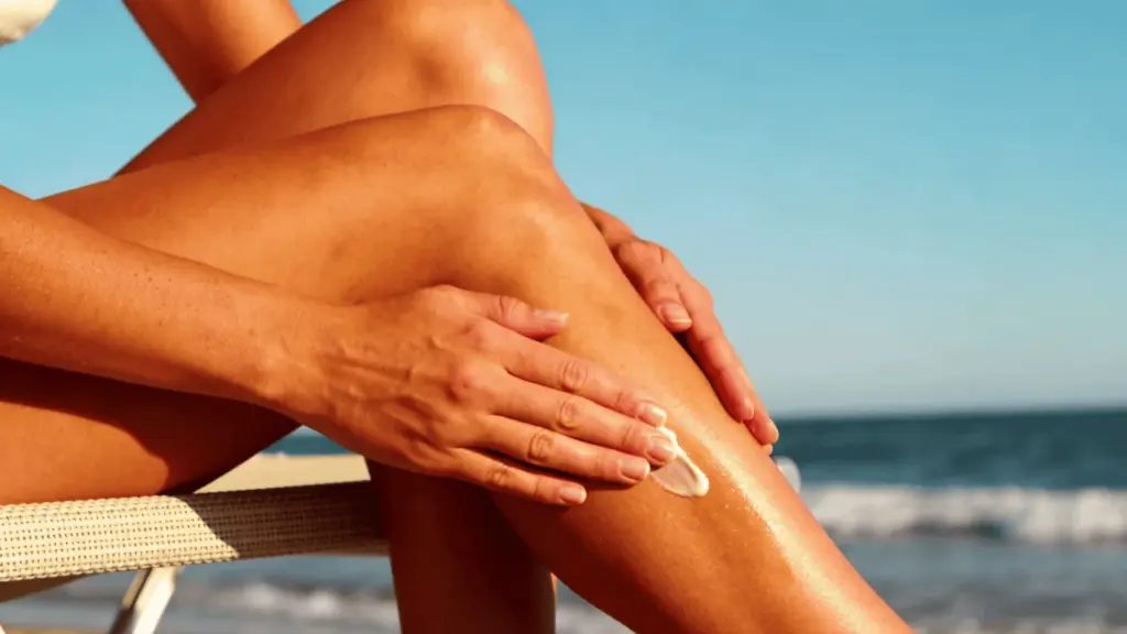 Close view of tanned legs on a beach as sunscreen is applied, showing even sun tan with ocean and blue sky in background