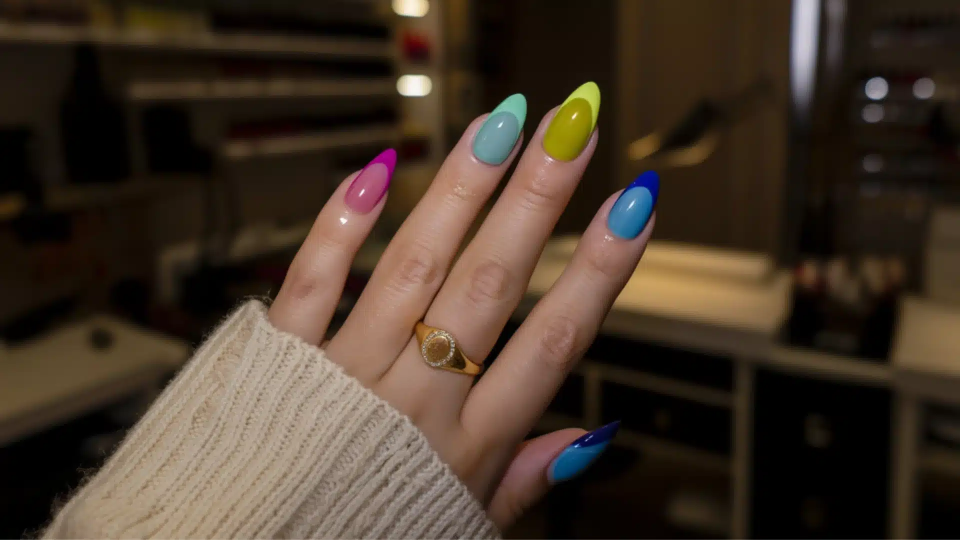 Colourful glossy almond-shaped nail art on feminine hand inside a dim luxury nail salon with warm ambient lighting and blurred background. (1)