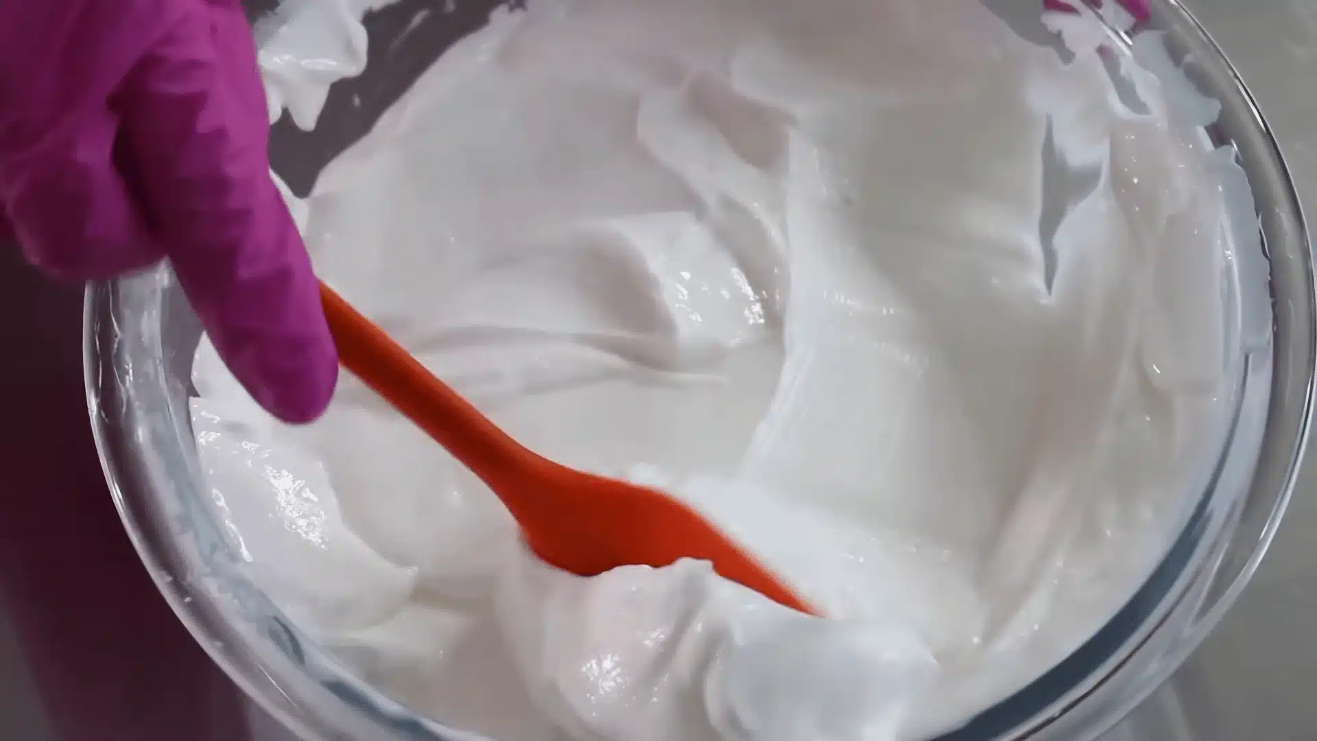 Creamy homemade body lotion being mixed in a glass bowl with a red spatula and gloved hand