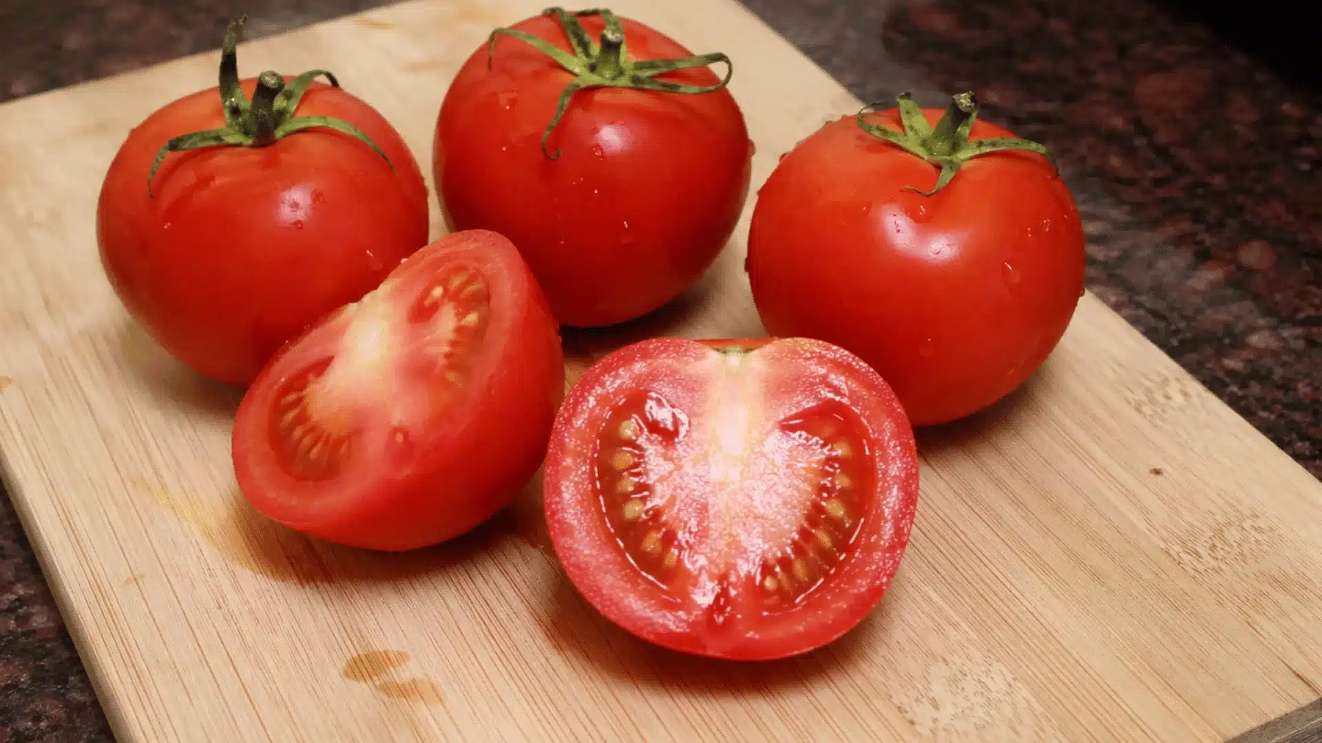 Fresh washed red tomatoes on a wooden chopping board, with whole tomatoes and a couple cut in half showing juicy interiors