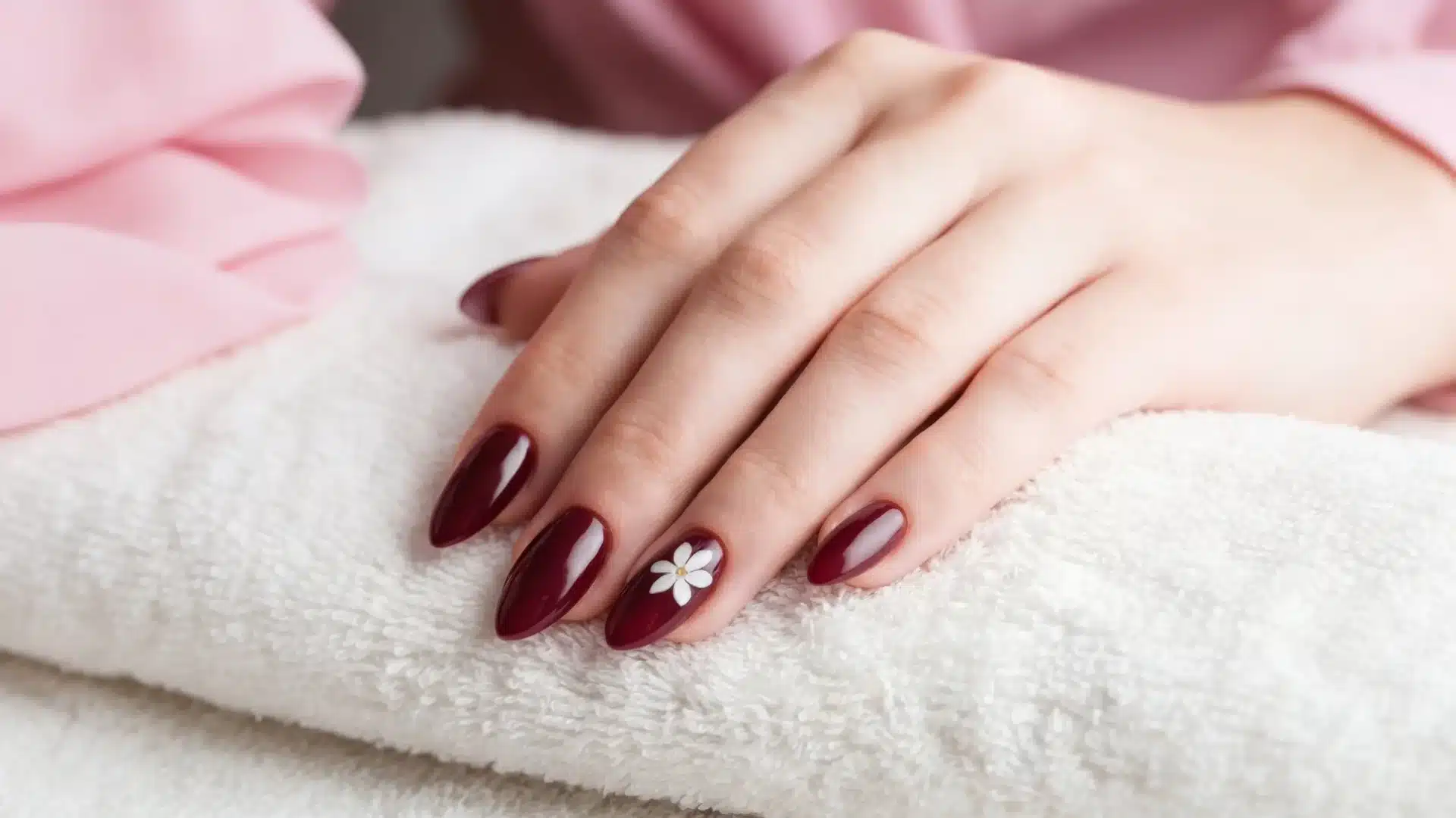 Hand with gel deep red gel nails featuring a white floral accent resting on a soft white towel