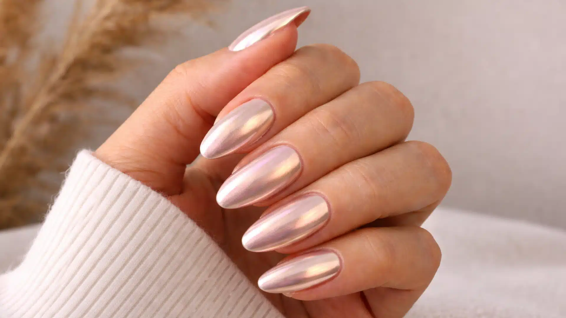 Hand with subtle chrome finish almond-shaped nails in soft pink tones, posed naturally with a cozy sweater against a warm neutral background