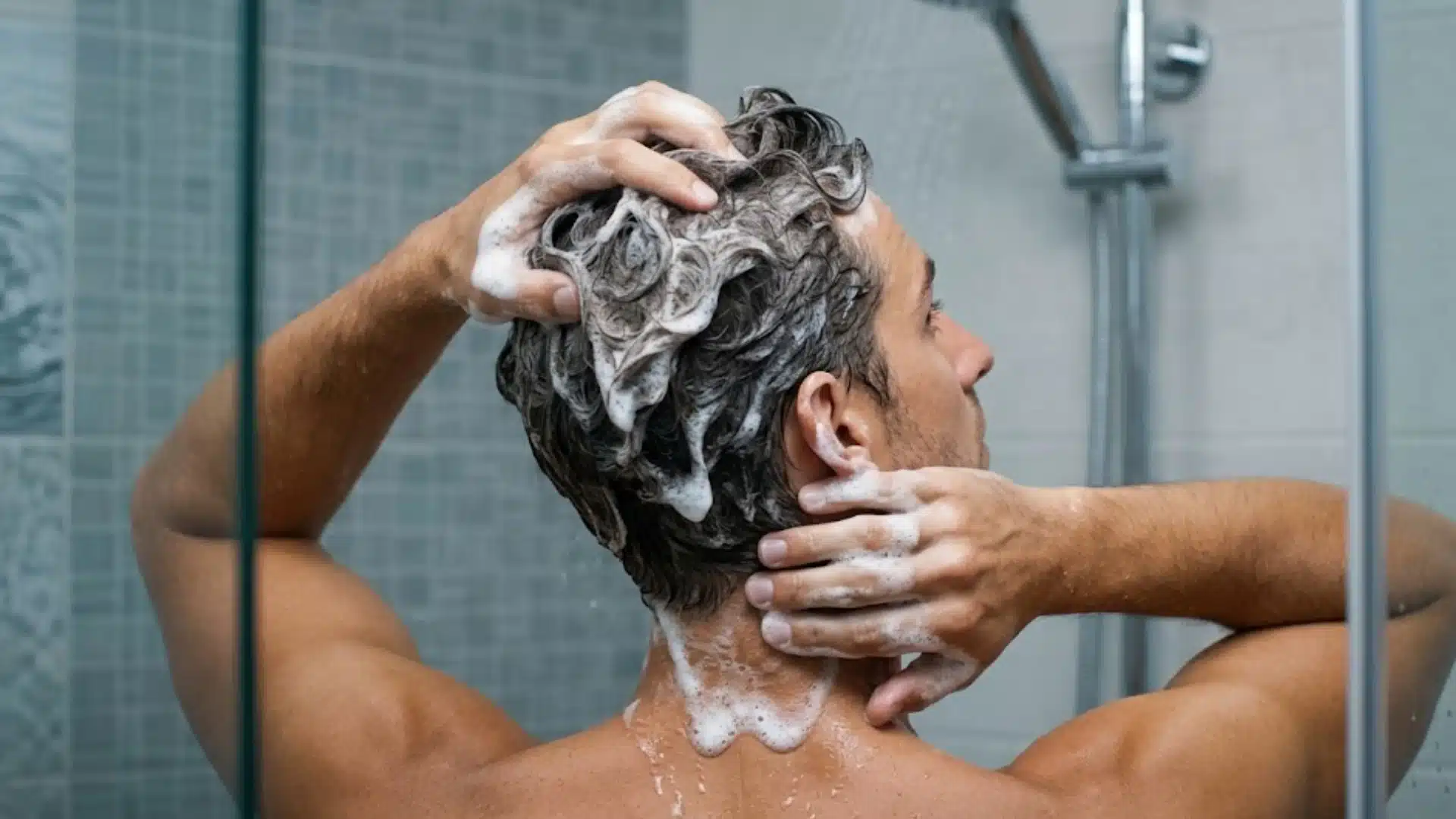 Man washing his hair in the shower with shampoo lather, seen from behind with hands on scalp