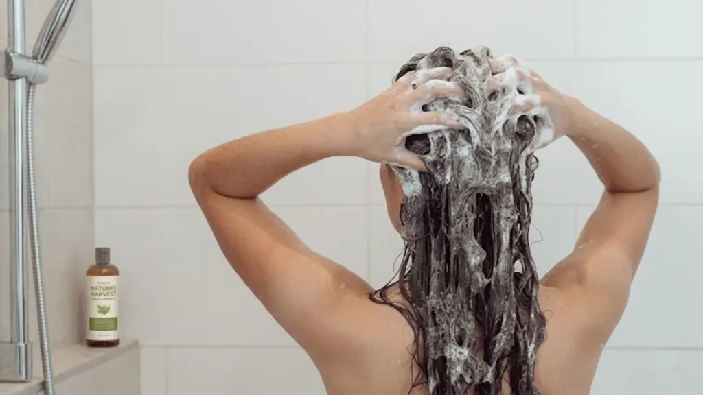 Person washing their long hair in the shower, hands massaging shampoo into scalp, viewed from behind