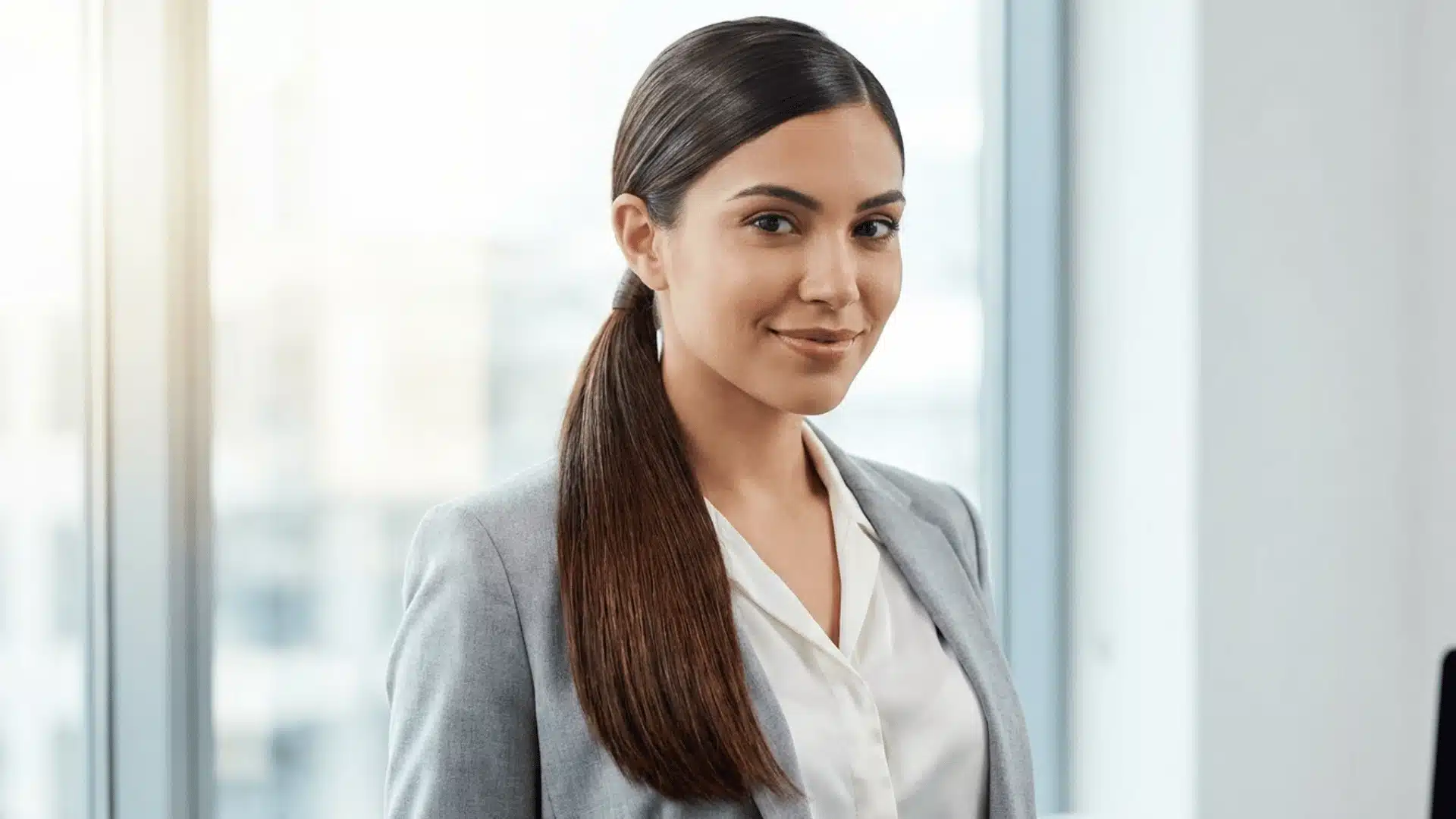 Professional woman in gray blazer smiling in modern office with sleek ponytail