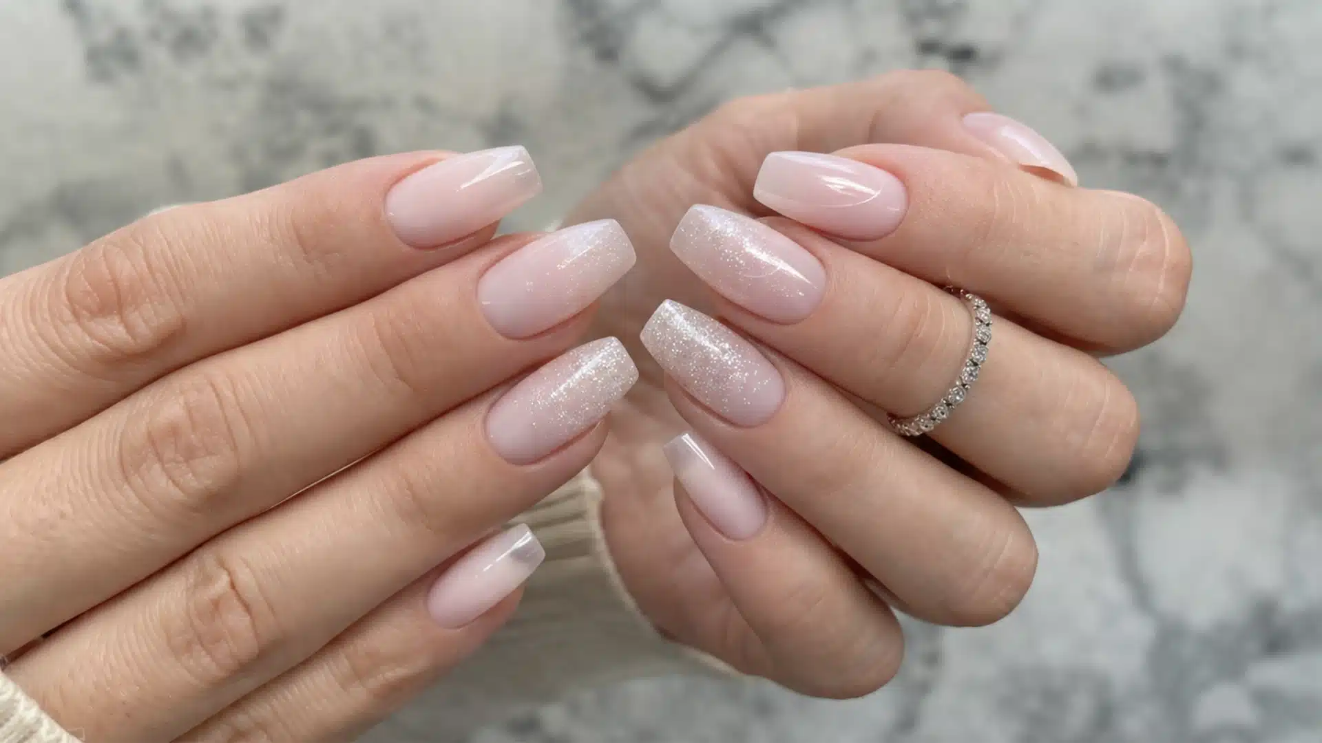 Soft pink ombre nails with subtle glitter accents and a silver ring, photographed in natural light with blurred outdoor background