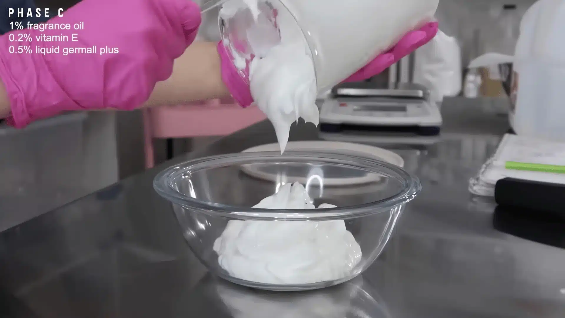 Thick homemade lotion being poured into a glass bowl by person wearing pink gloves
