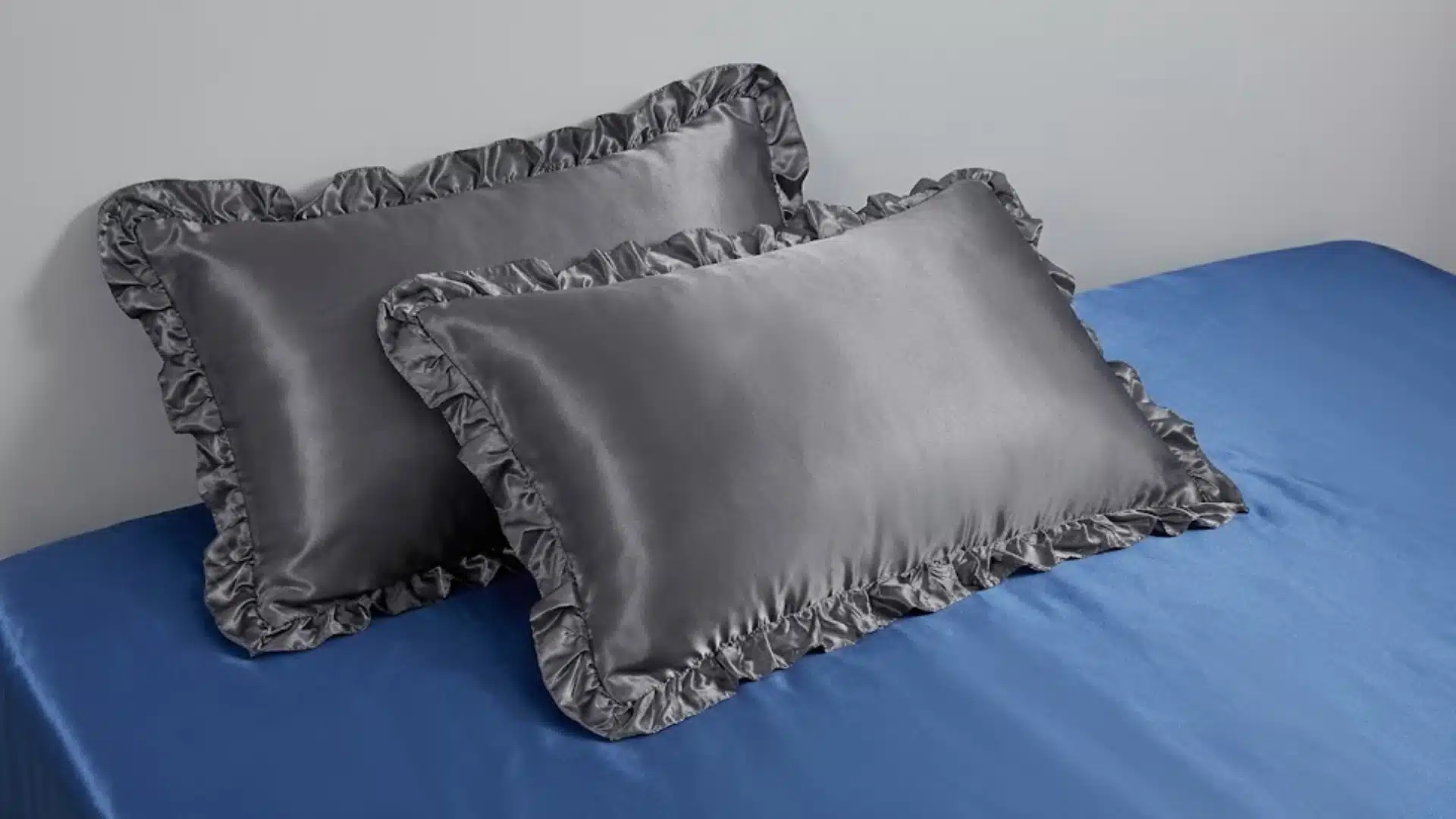 Two gray satin pillowcases with ruffled edges placed on blue bed, showing smooth texture and soft sheen