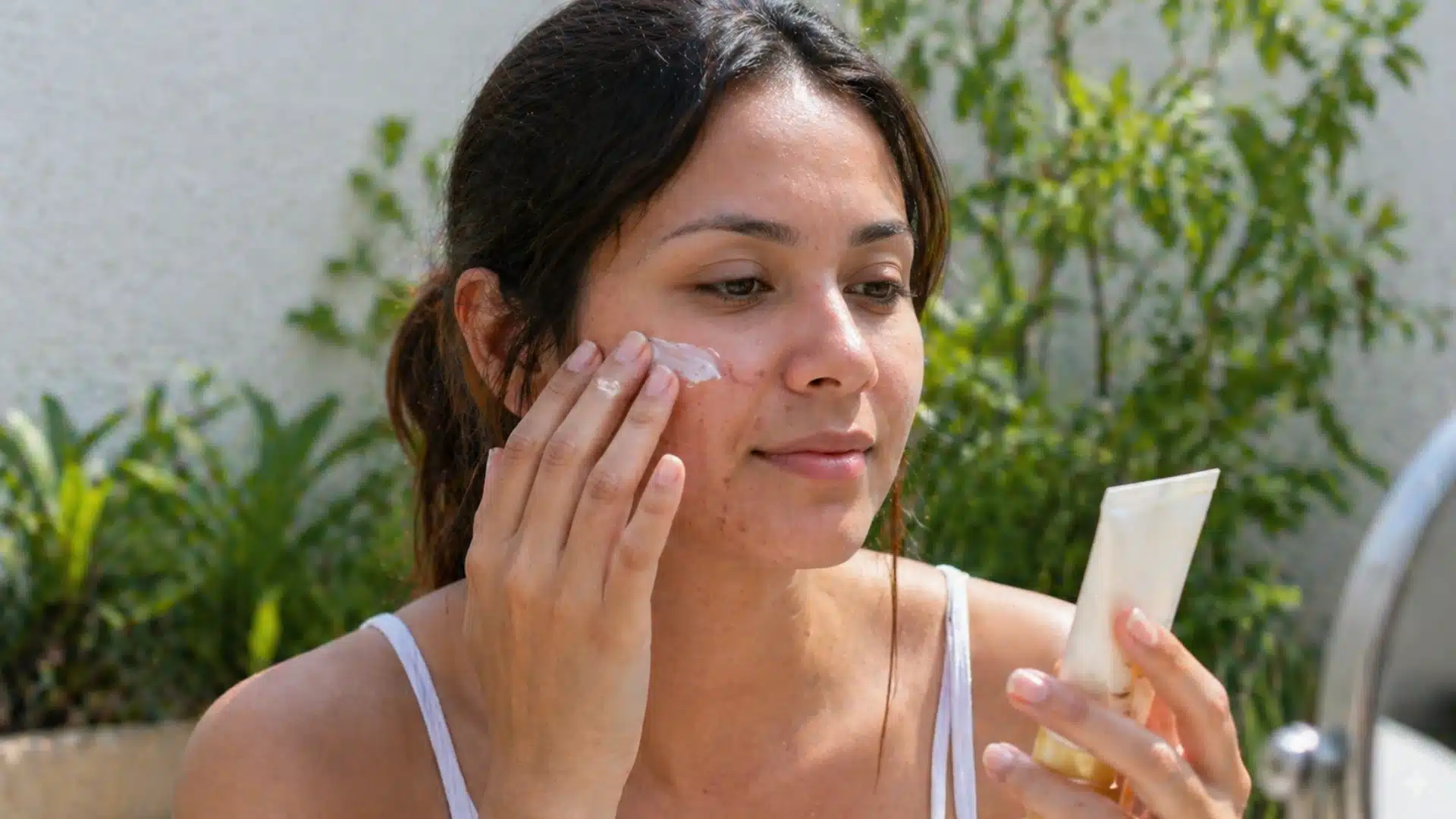 Woman applying acne-safe sunscreen on face outdoors, holding SPF 50 tube, natural skin texture visible in soft daylight setting