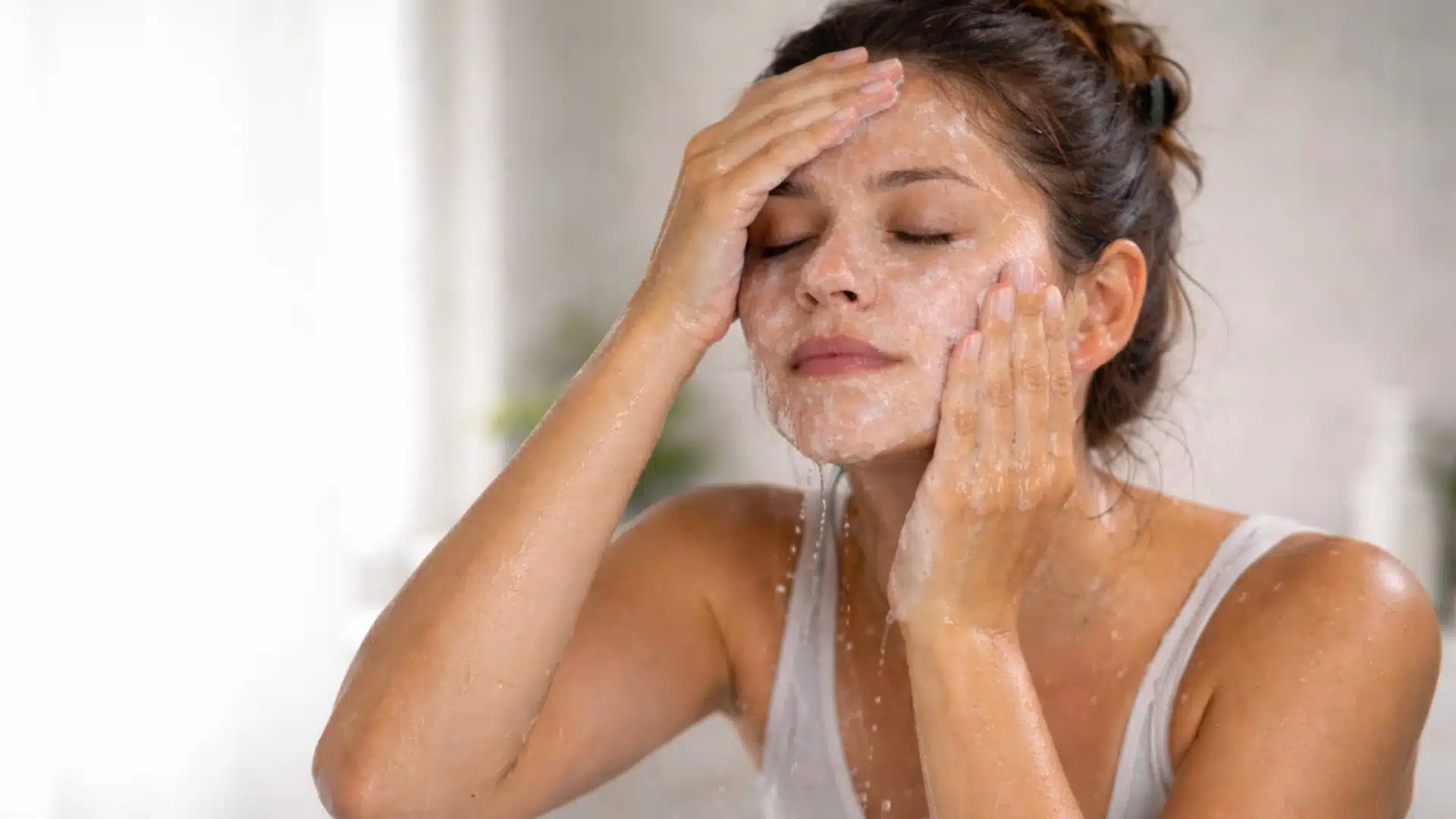 Woman washing her face with a gentle cleanser, massaging foam into skin in a bright bathroom setting with water droplets visible
