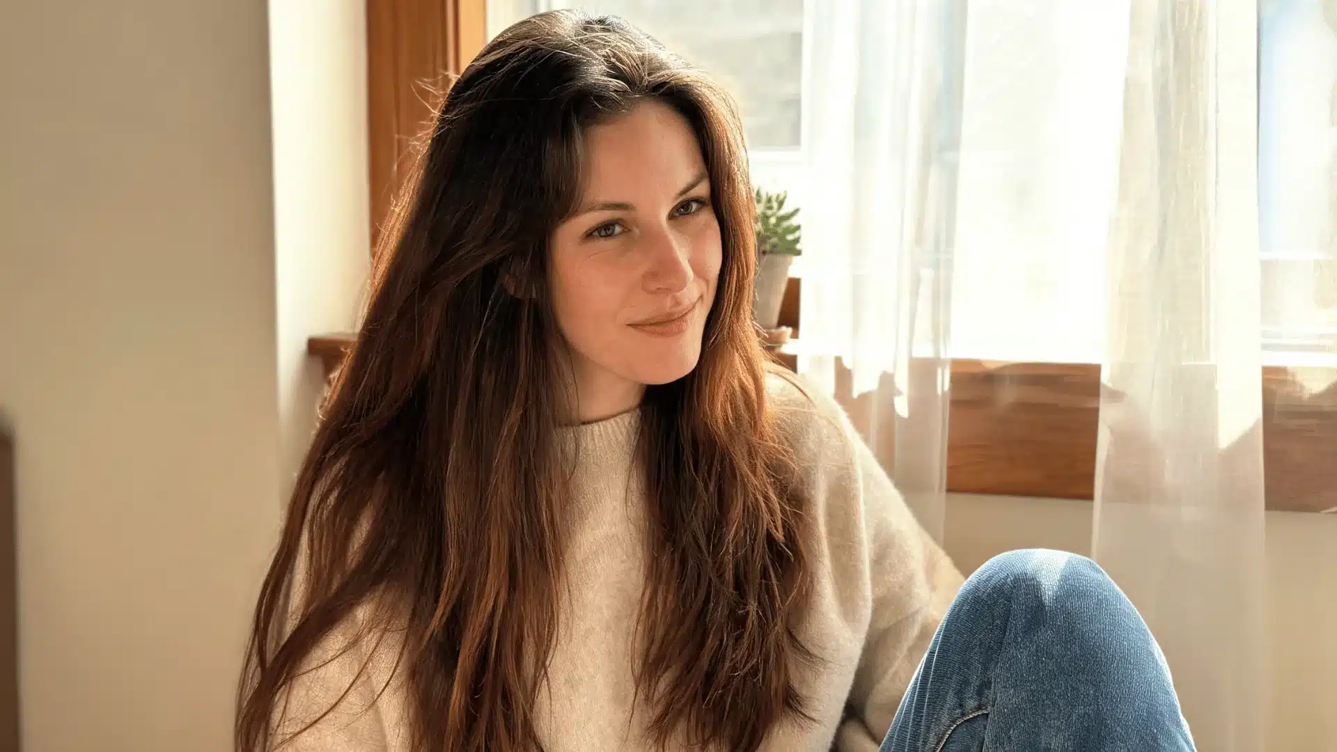 Woman with long brown hair sitting by a sunlit window in a cozy room