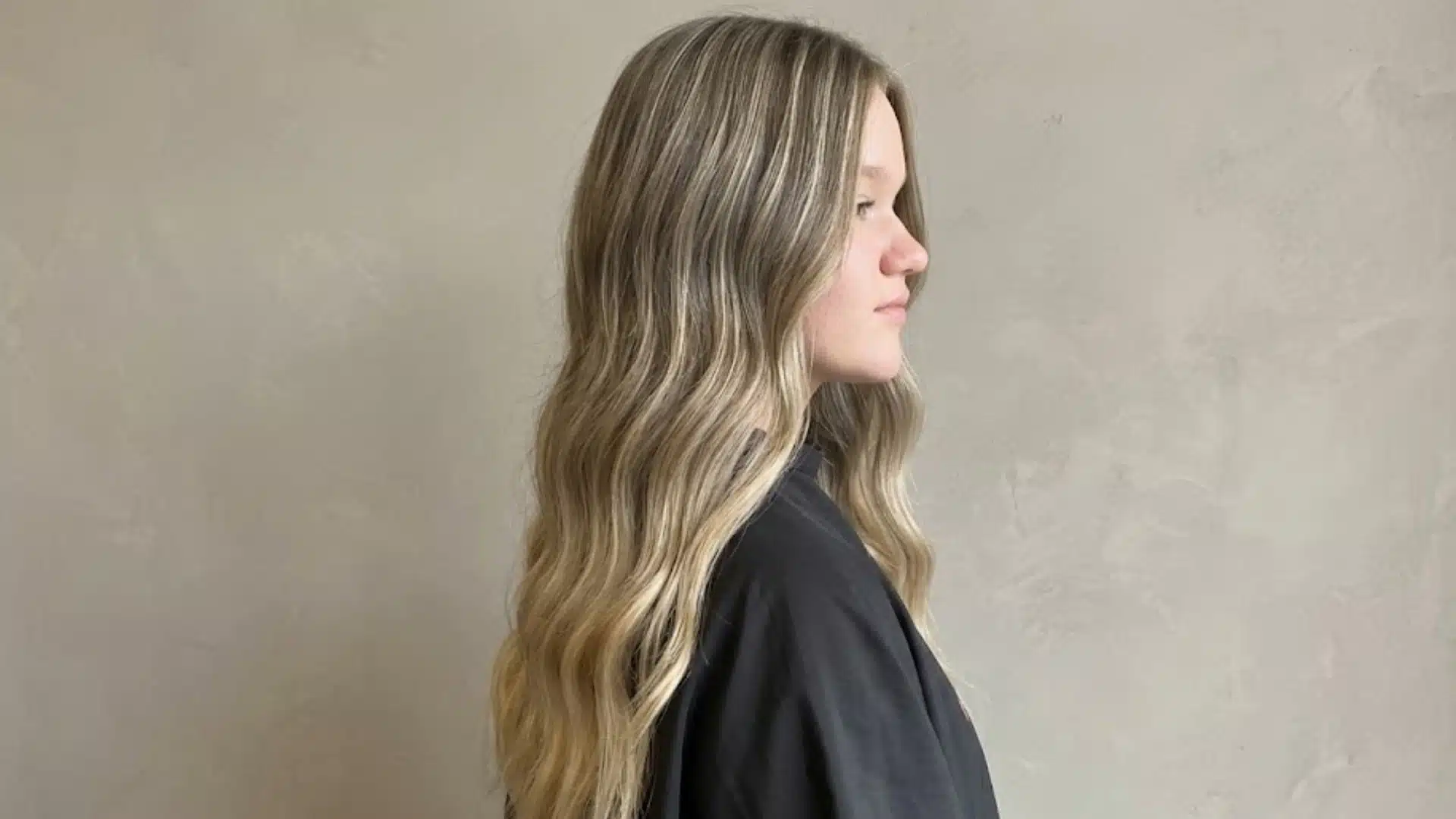 Woman with long soft blonde balayage waves and layered hairstyle shown from side profile indoors