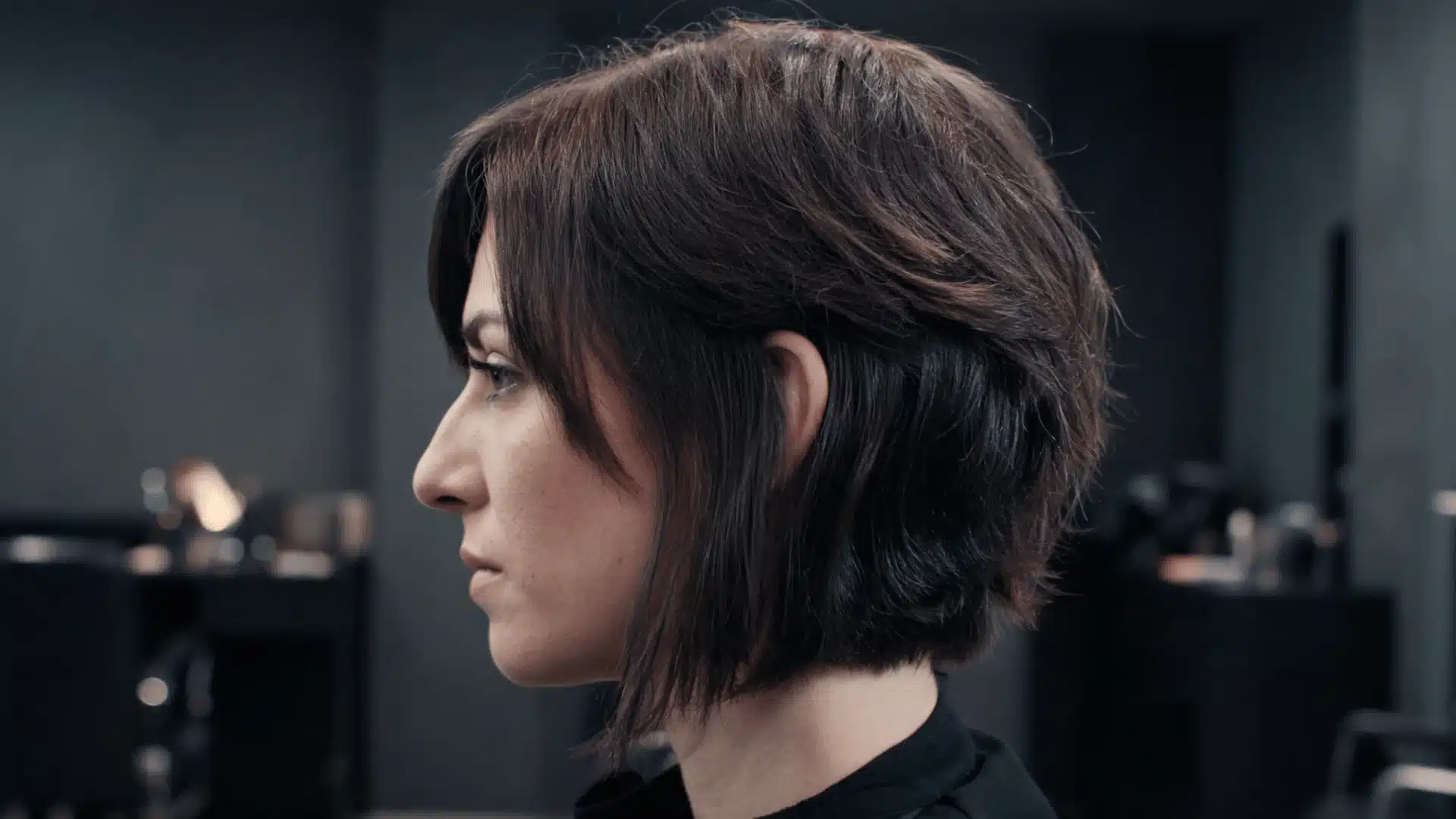 Woman with short dark layered hair shown in side profile against a dark salon background