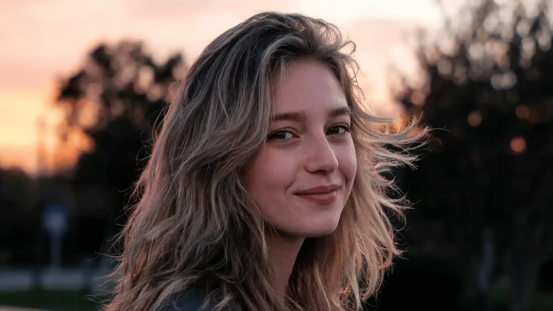 Woman with wavy blonde hair smiling outdoors during sunset with trees in the background