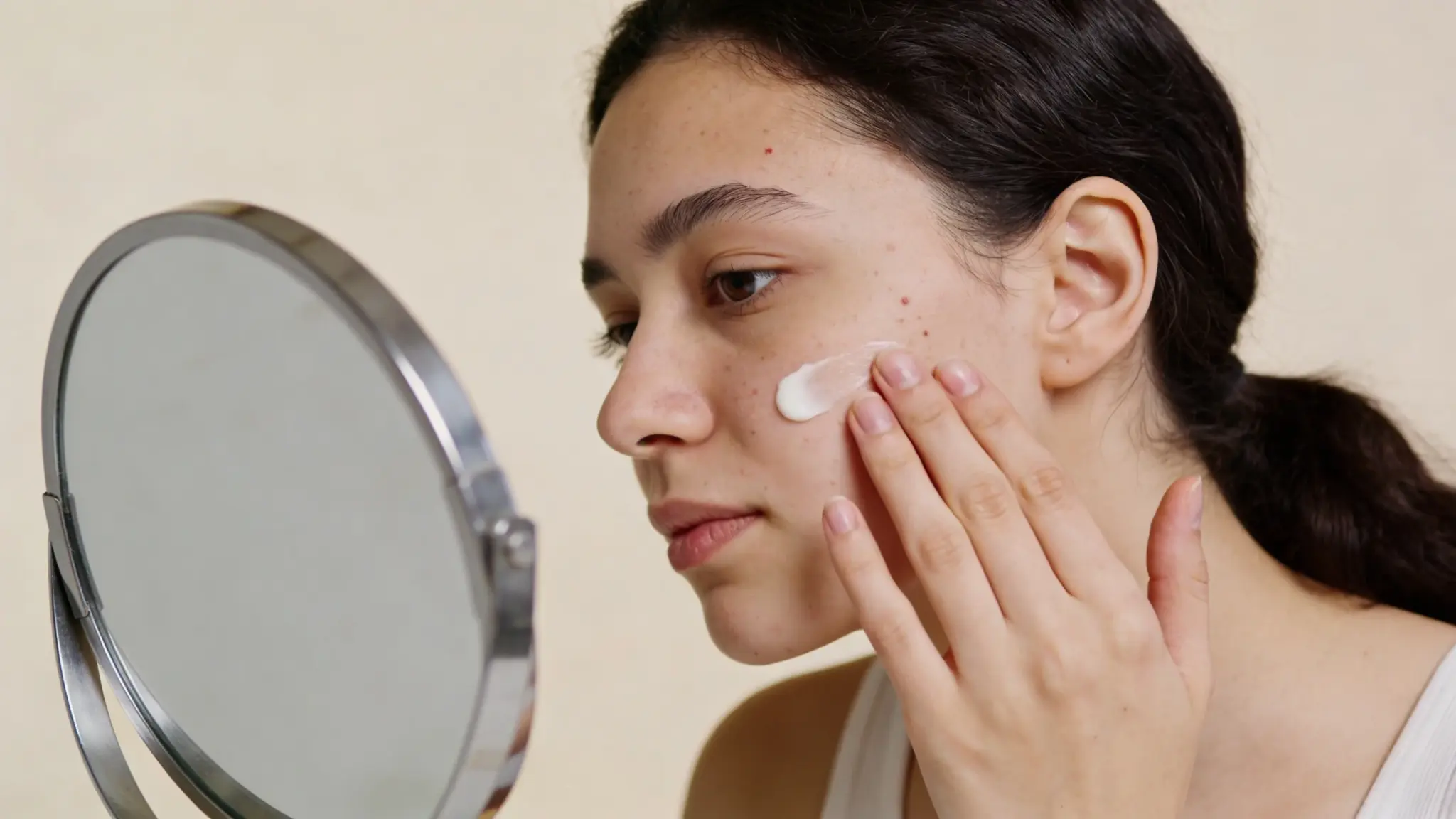 Young woman applying facial cream to cheek while looking in mirror, close up skincare routine at home