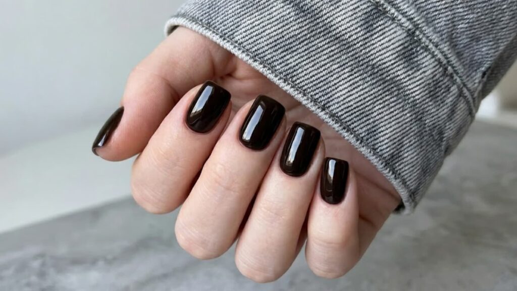 A fair-skinned hand showing glossy, deep dark brown square nails with a grey denim jacket cuff