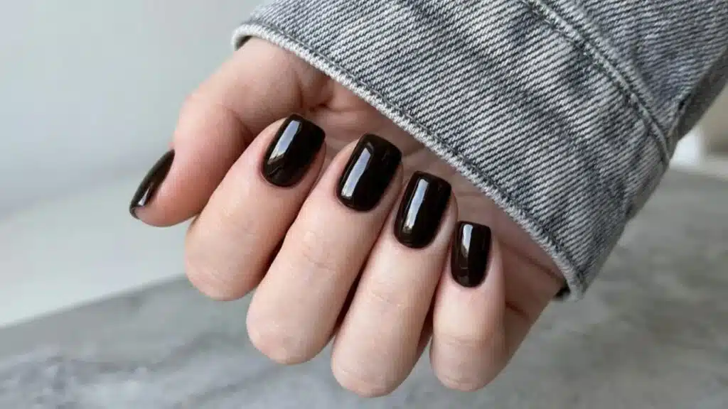 A fair-skinned hand showing glossy, deep dark brown square nails with a grey denim jacket cuff