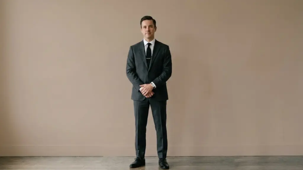 A man in a dark grey suit, white shirt, and black tie stands with hands clasped against a plain beige wall.
