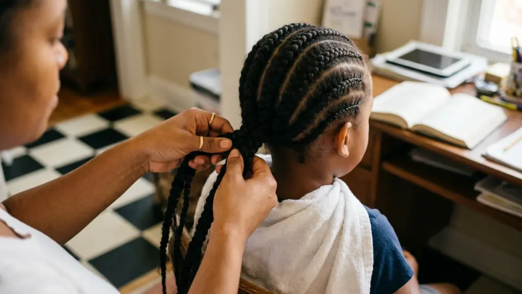 A person braids a child’s hair in neat cornrows while the child sits near a desk with an open book.