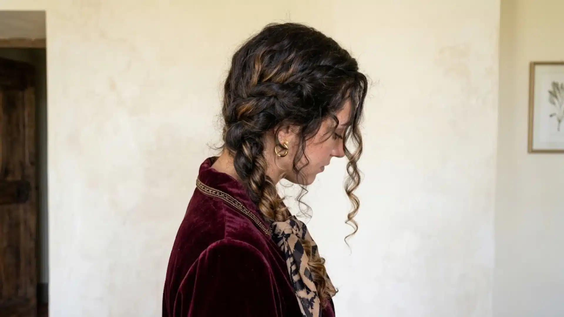 A side profile of a woman with braided curly hair wearing a maroon velvet blazer in a rustic room