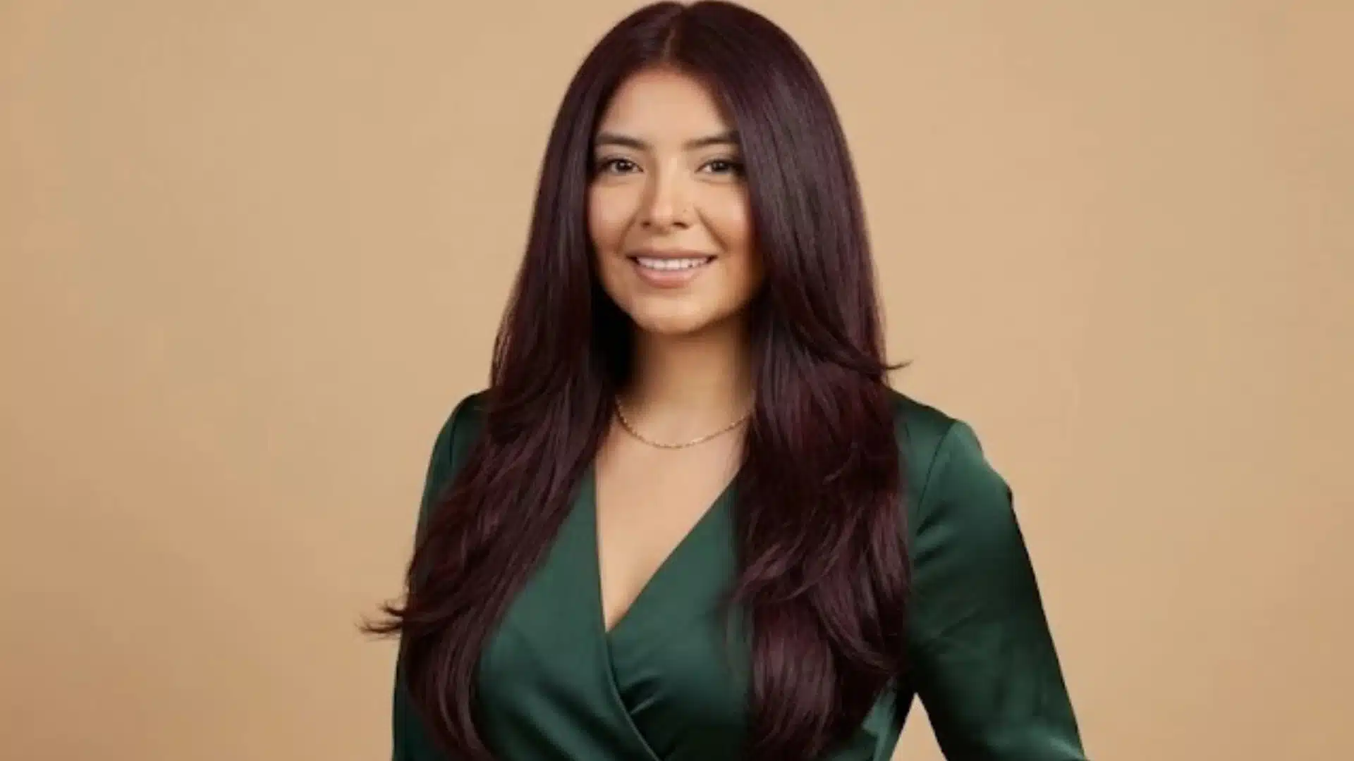 A smiling woman with cool mahogany hair in a forest green wrap dress with a hand in her pocket, against a solid tan background