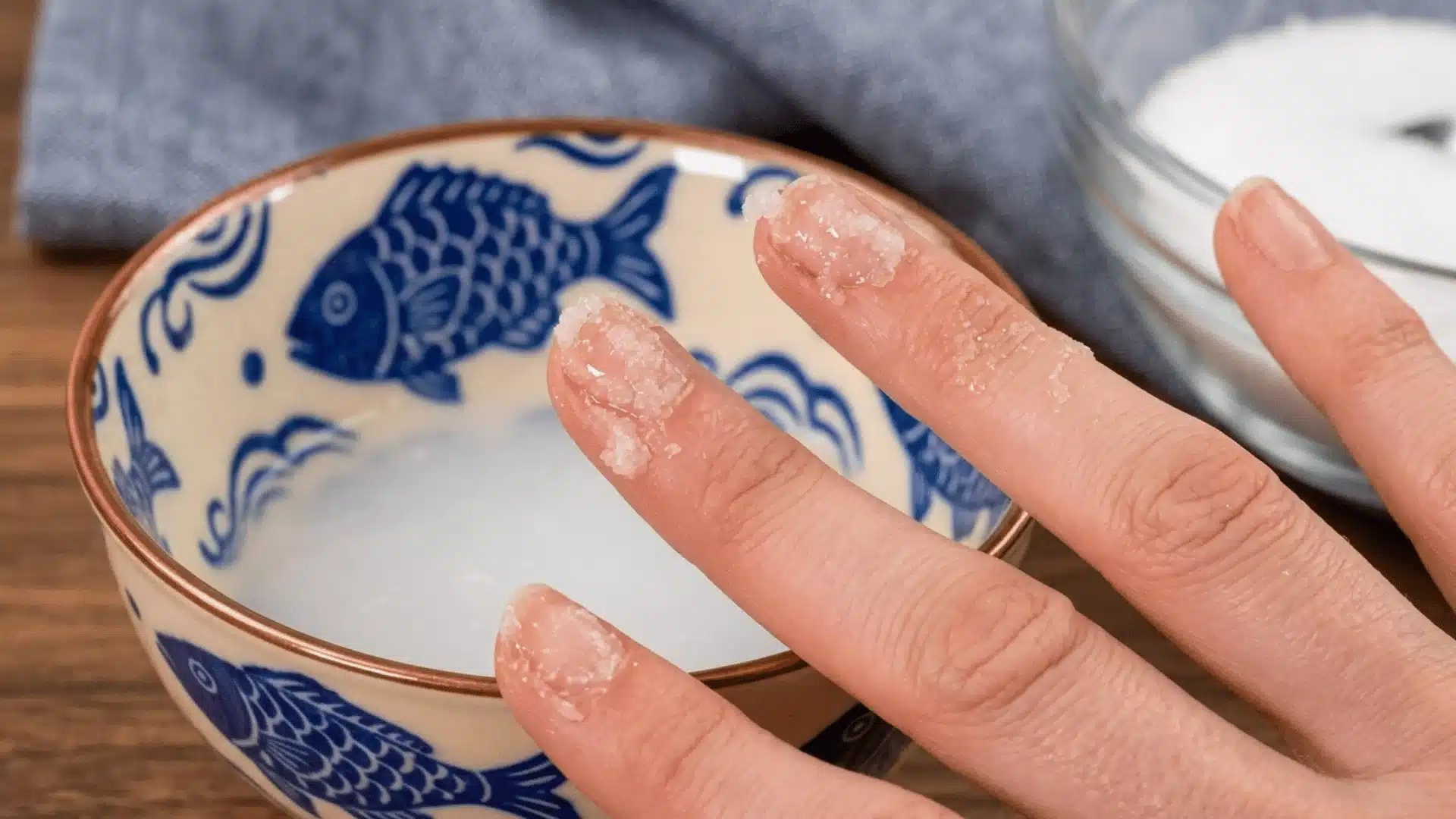 A close-up of fingers with coarse coconut oil scrub over a blue-and-white fish pattern bowl, on a wooden table