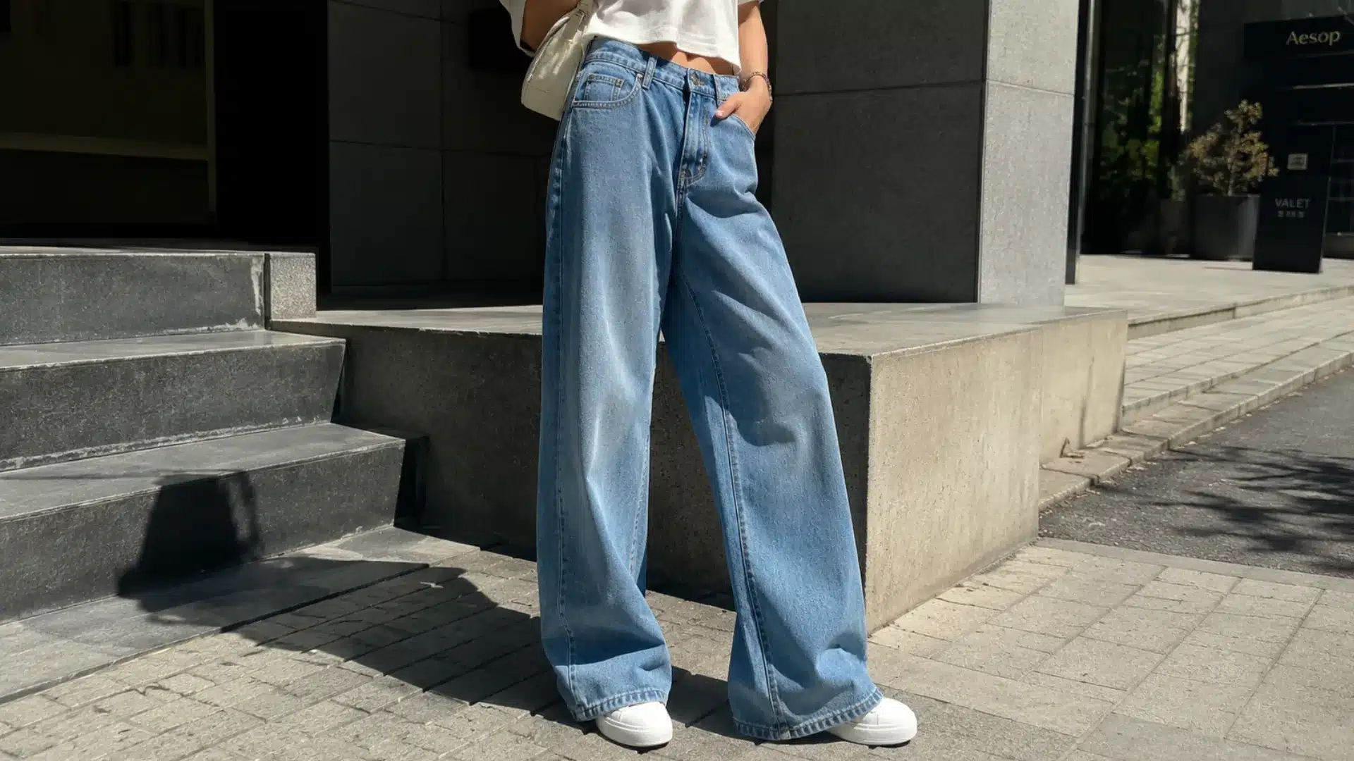 A person wearing wide-leg light wash jeans, a white crop top, and white sneakers standing on a city sidewalk